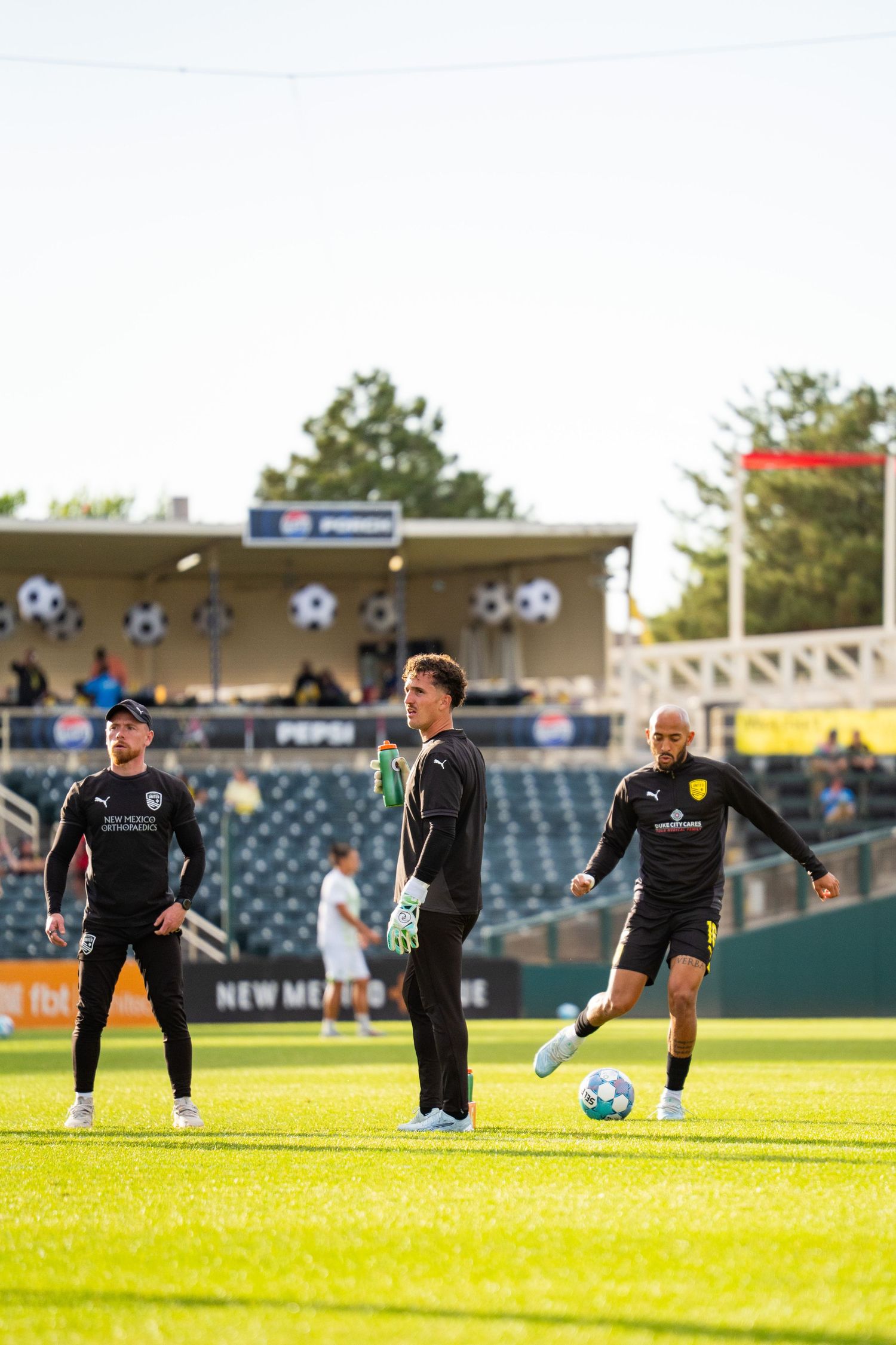 Soccer players in black uniforms warming up on a green field at a stadium during a sunny evening match.