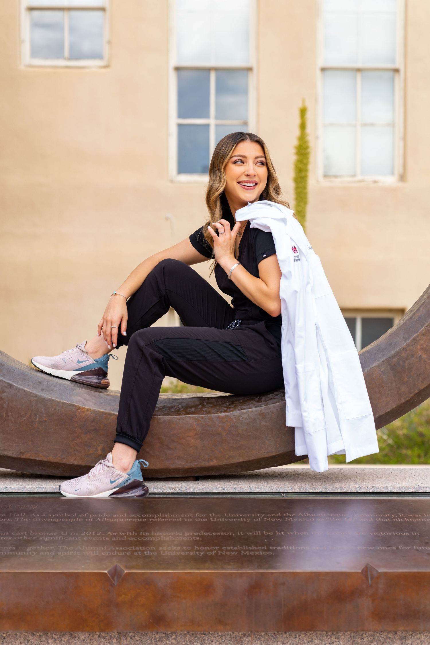 Healthcare worker in scrubs and white coat sitting on metal sculpture outside medical building.