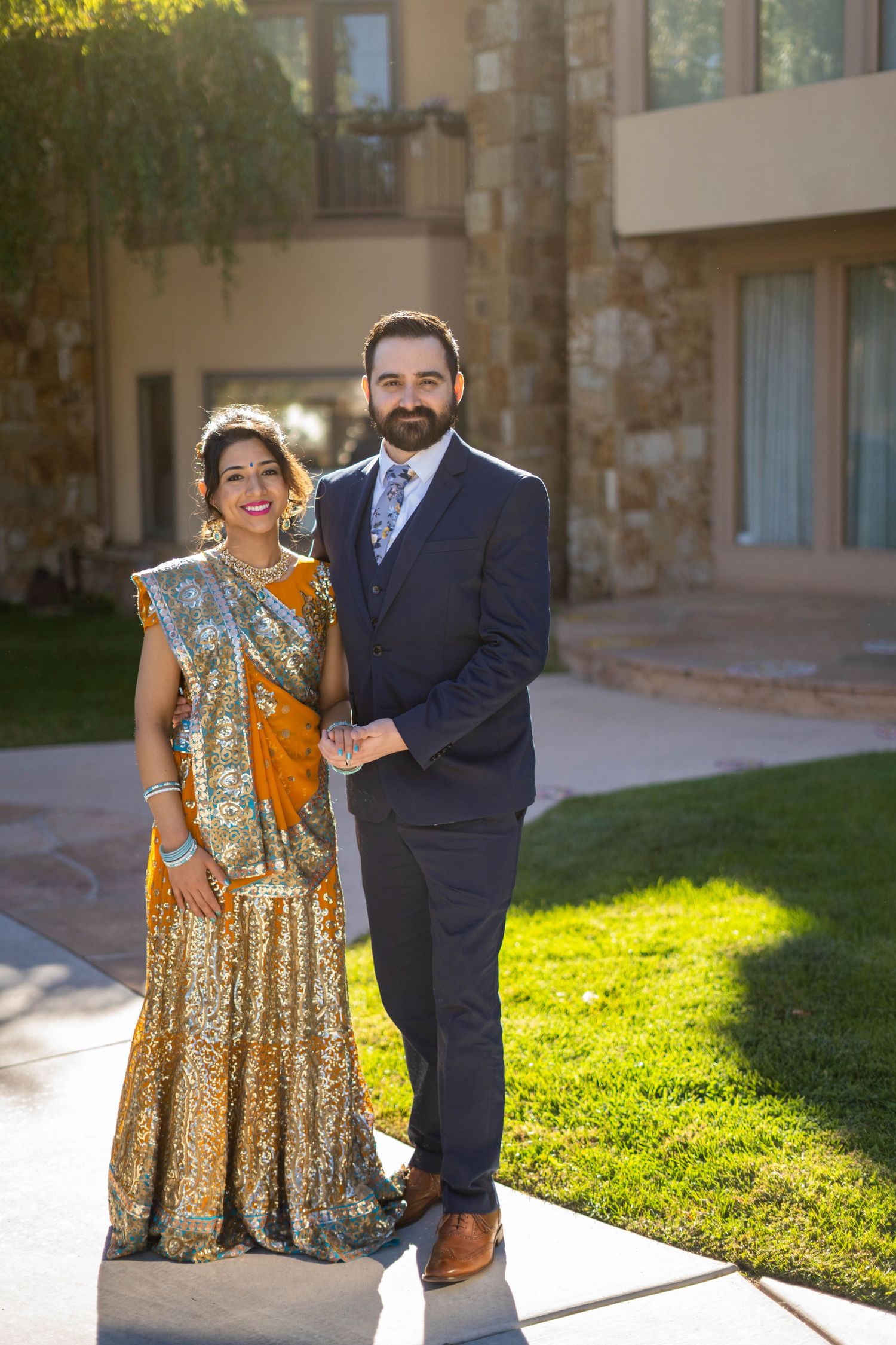 A couple in traditional Indian attire poses outdoors, with the woman wearing a sparkling gold lehenga against green landscaping.