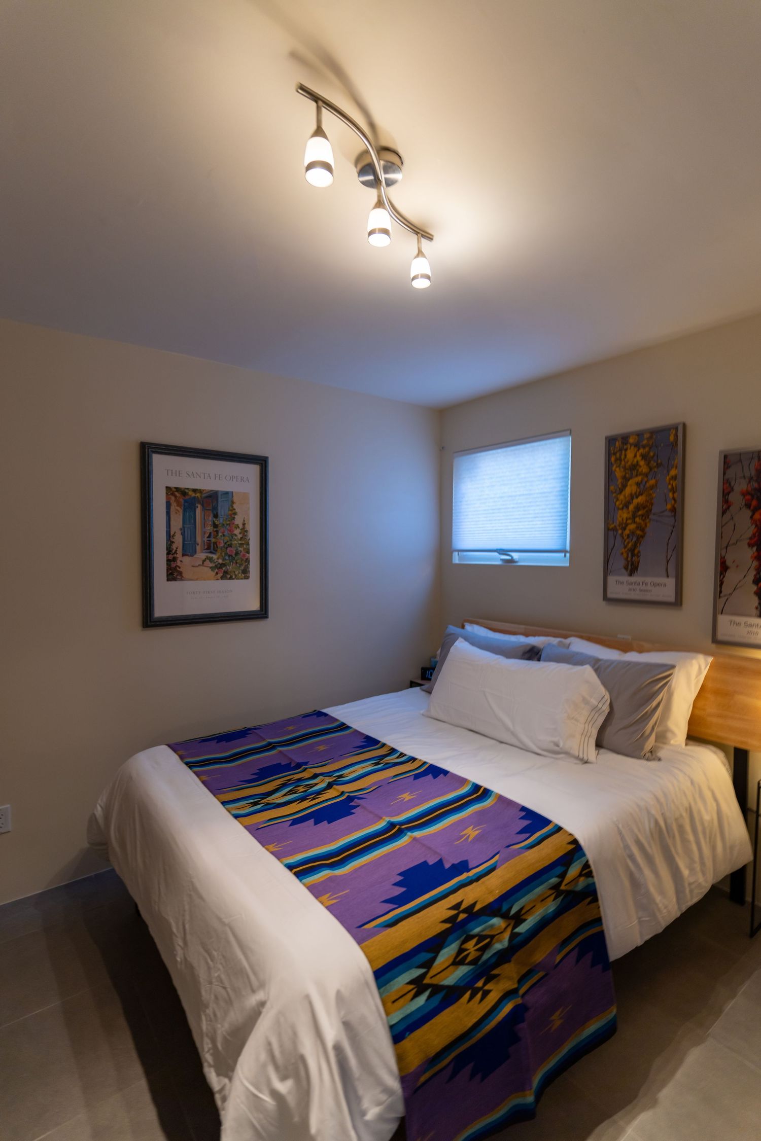 Modern bedroom interior with white bedding and colorful geometric patterned blanket under track lighting on ceiling.