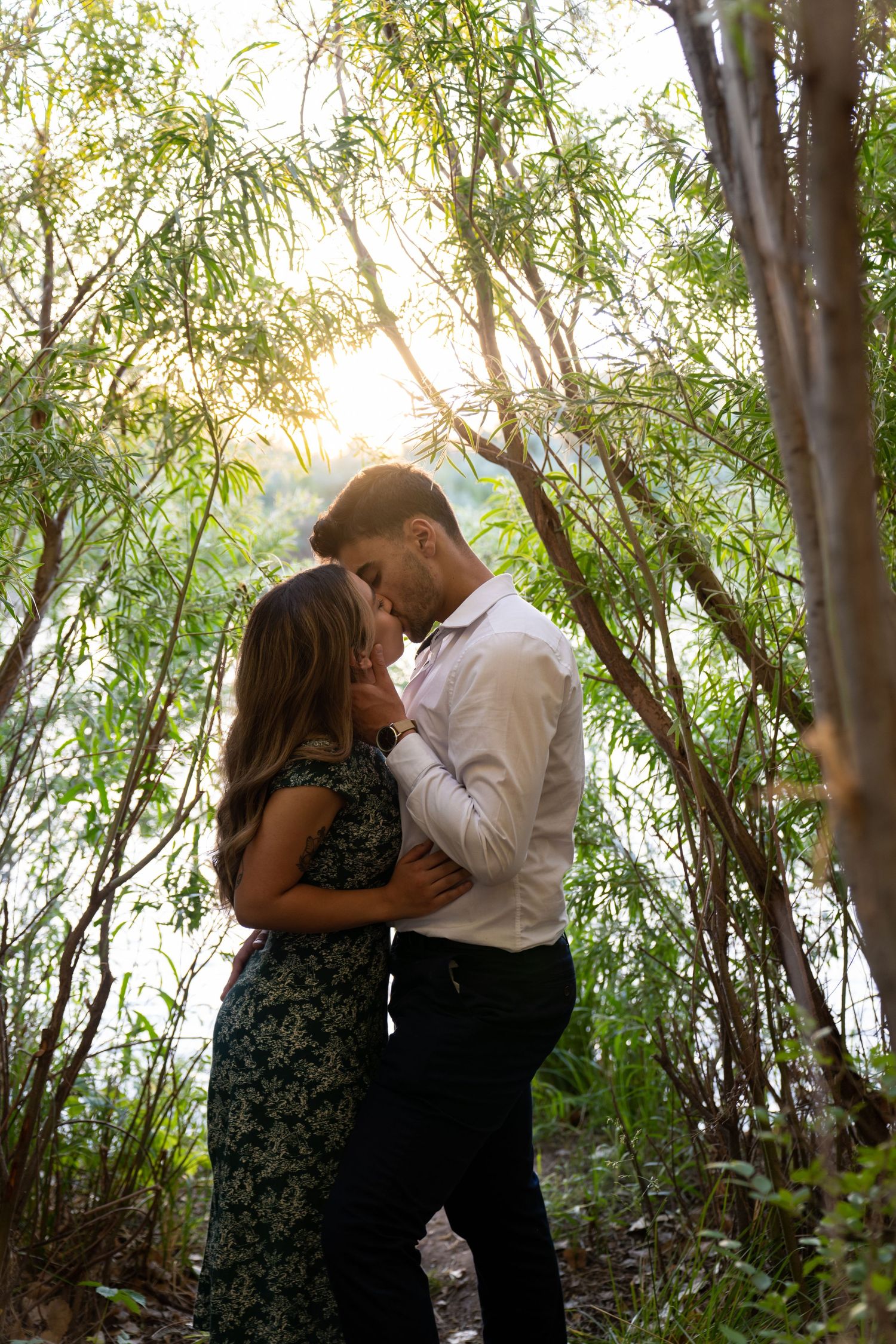 A romantic couple shares intimate moments in a wooded setting with willow trees during golden hour.