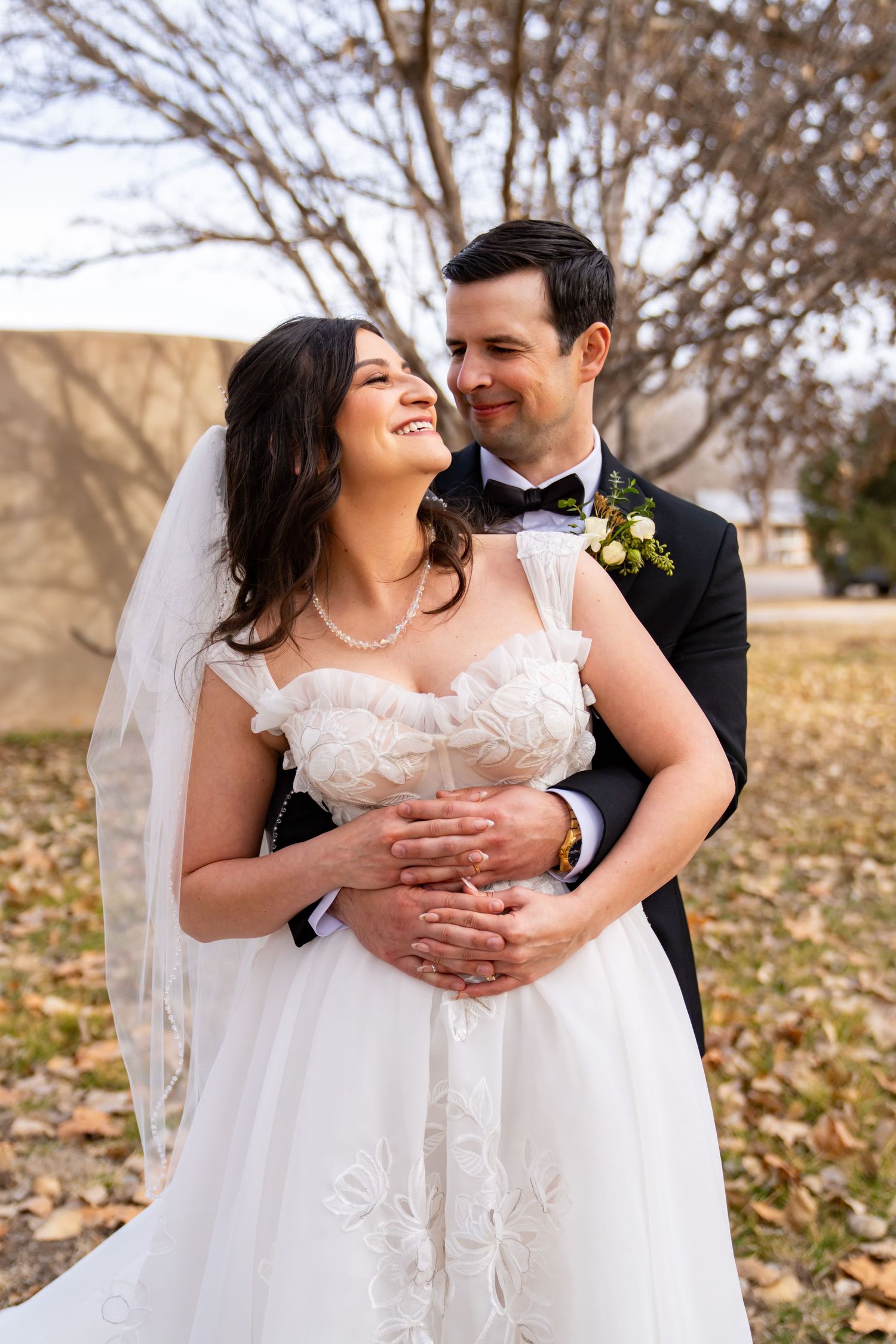A smiling bride in a white gown and veil embraces her groom in a black tuxedo outdoors on their wedding day.