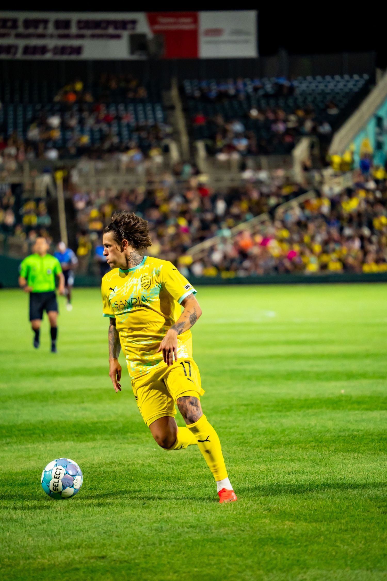 Soccer player in yellow jersey #17 dribbles ball on green field during night match with stadium crowd in background.