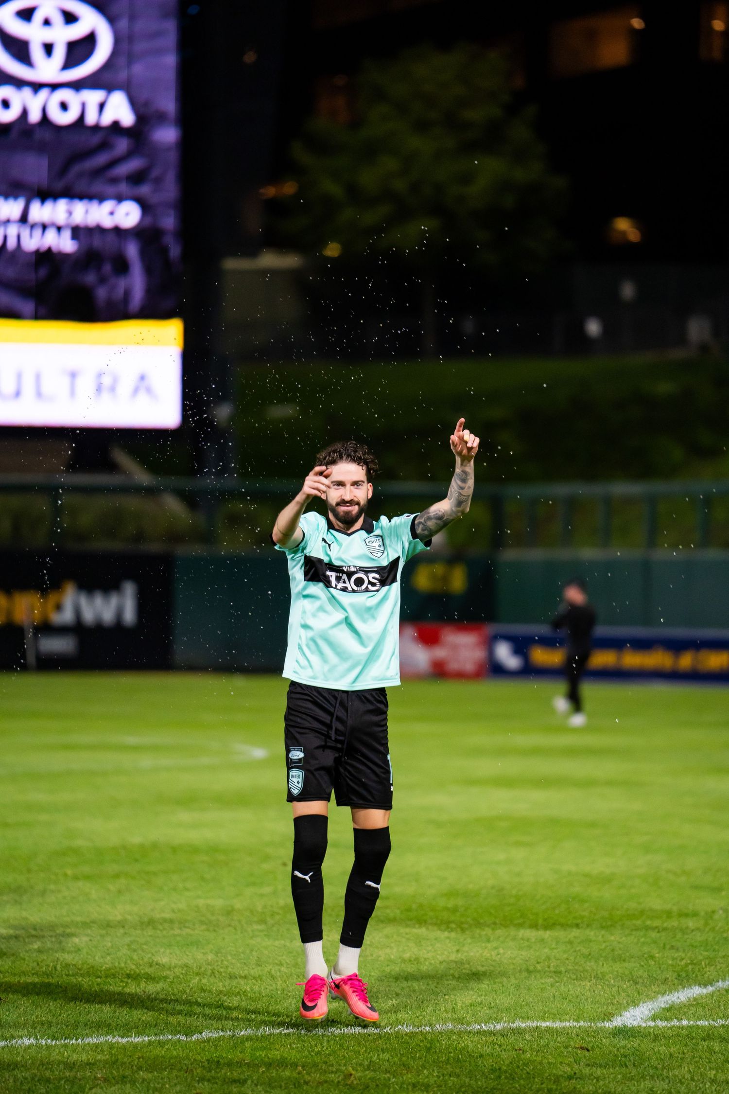 Soccer player in teal New Mexico United jersey clapping on green field at night stadium with Toyota signage.