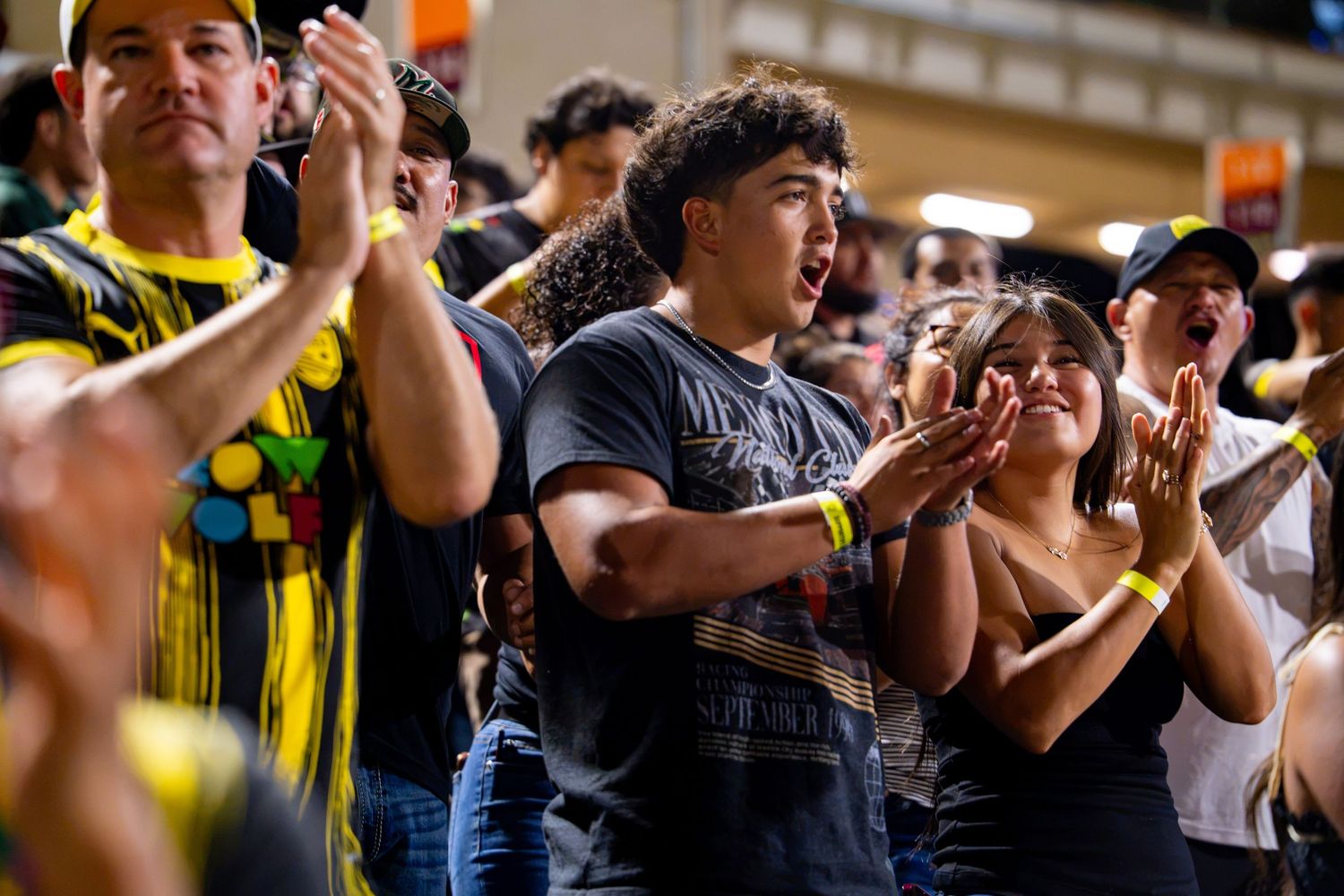 Excited sports fans clapping and cheering in stadium stands, wearing casual attire at a live event.