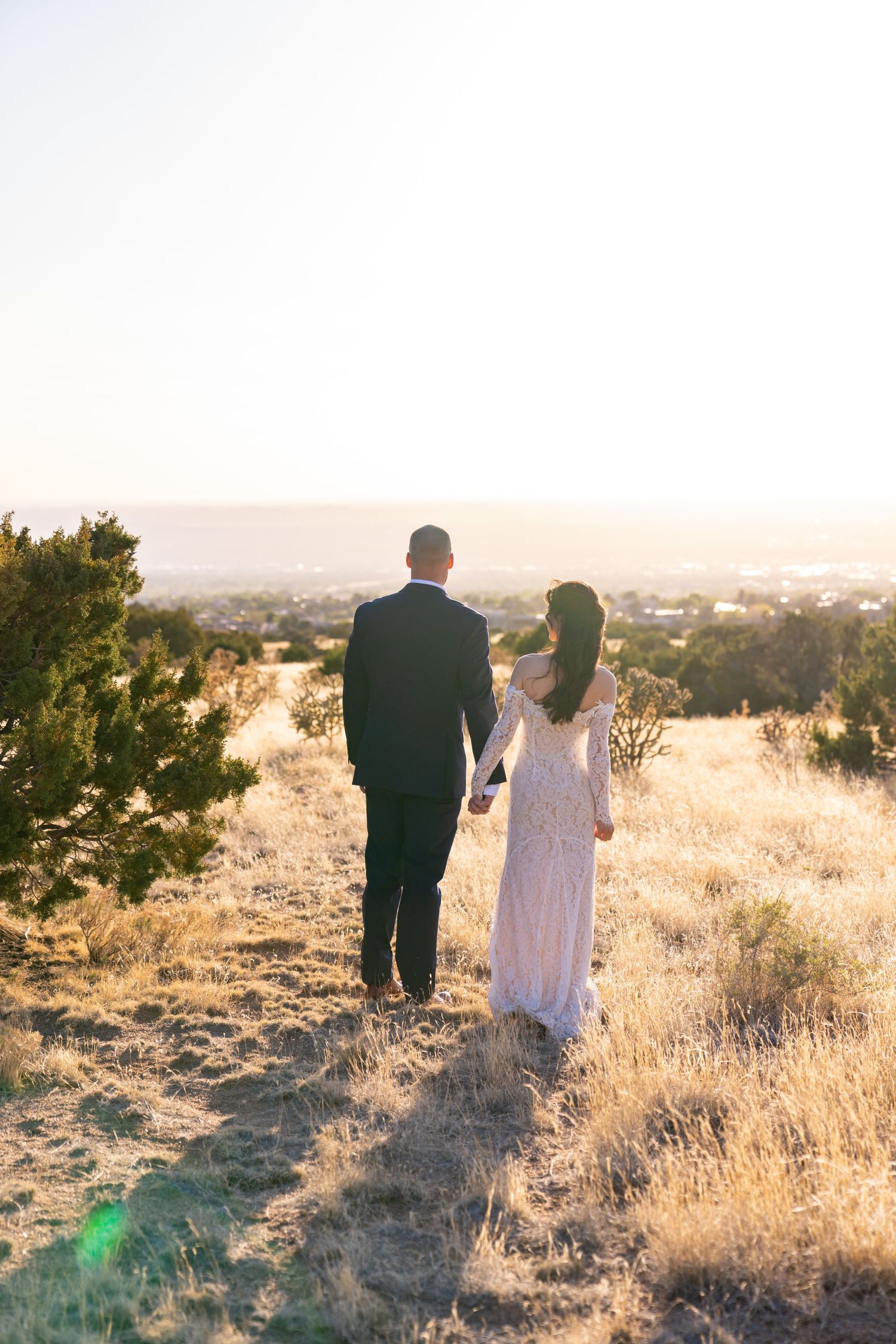 A couple in wedding attire holds hands walking through golden fields at sunset with a scenic valley view.
