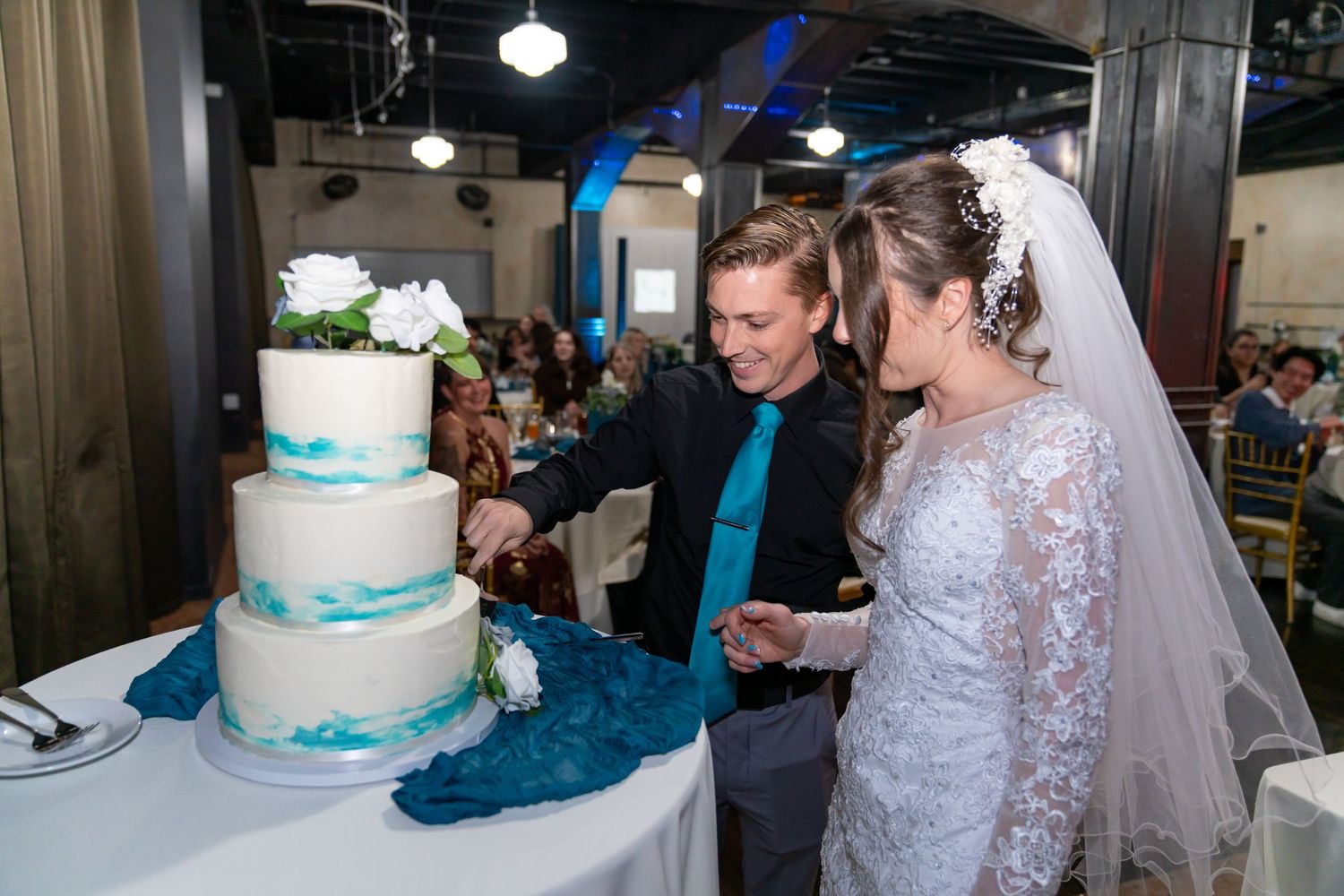 Bride and groom cutting a three-tier white wedding cake with teal accents at their wedding reception.