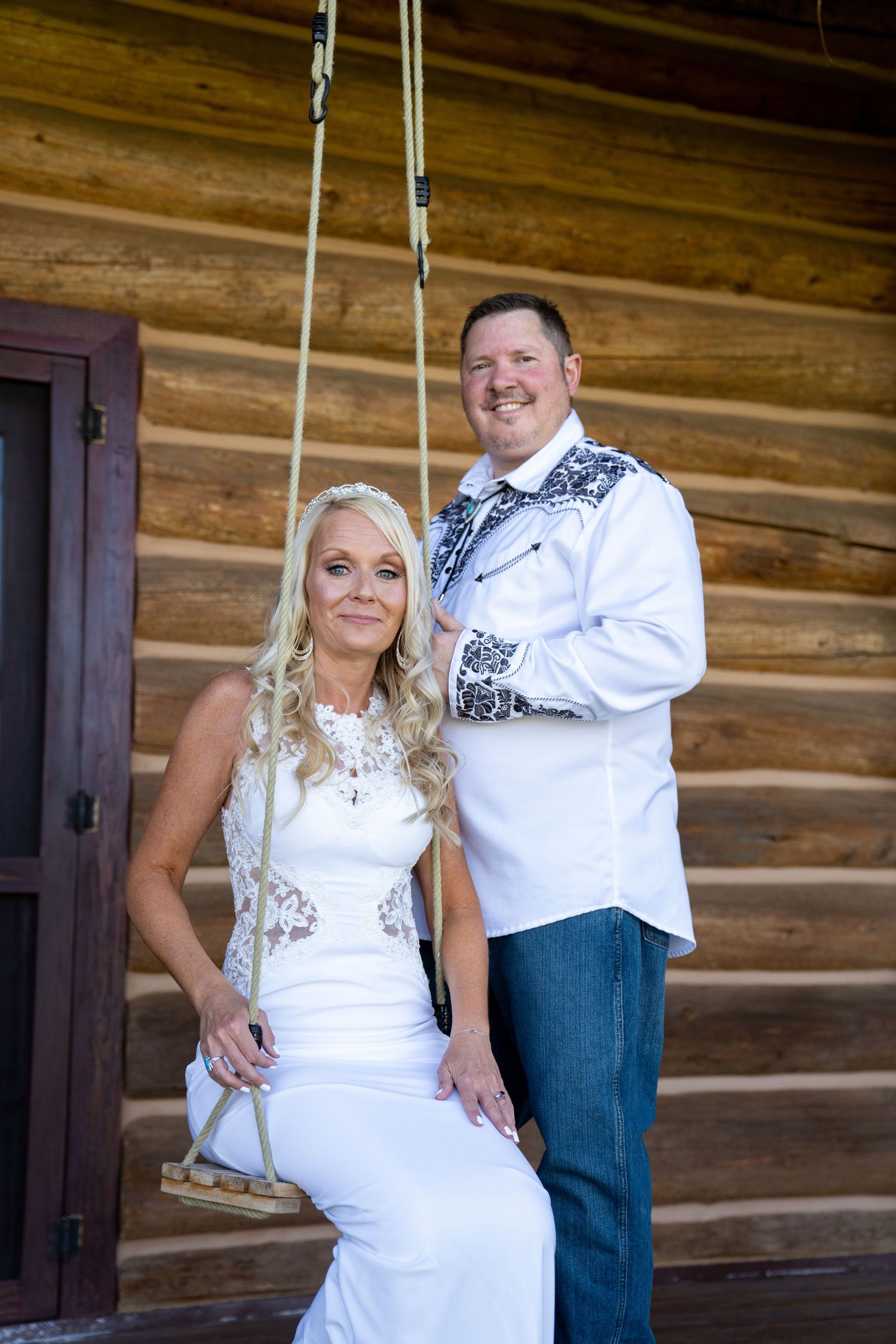 Couple in rustic wedding attire pose playfully against a log cabin exterior wall.