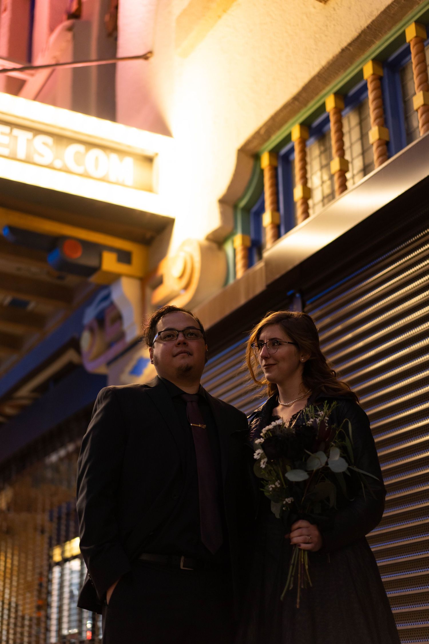 A couple stands together in front of a storefront at night with warm lighting creating an intimate atmosphere.