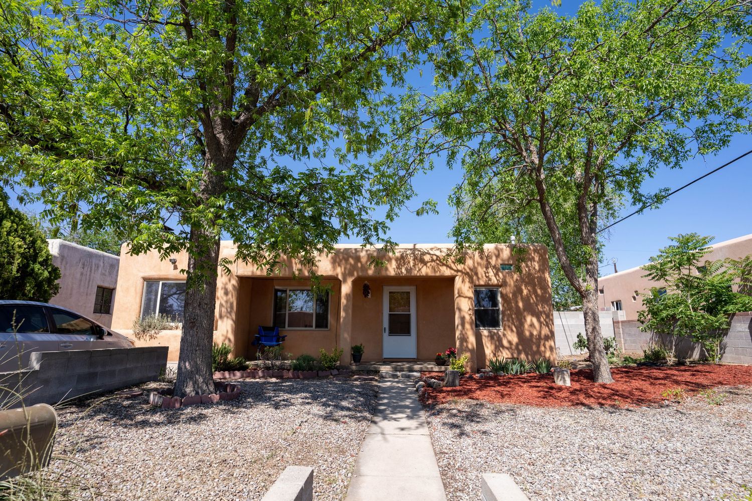 A panoramic view of a desert landscaped backyard with gravel, trees, and adobe style buildings in the background.