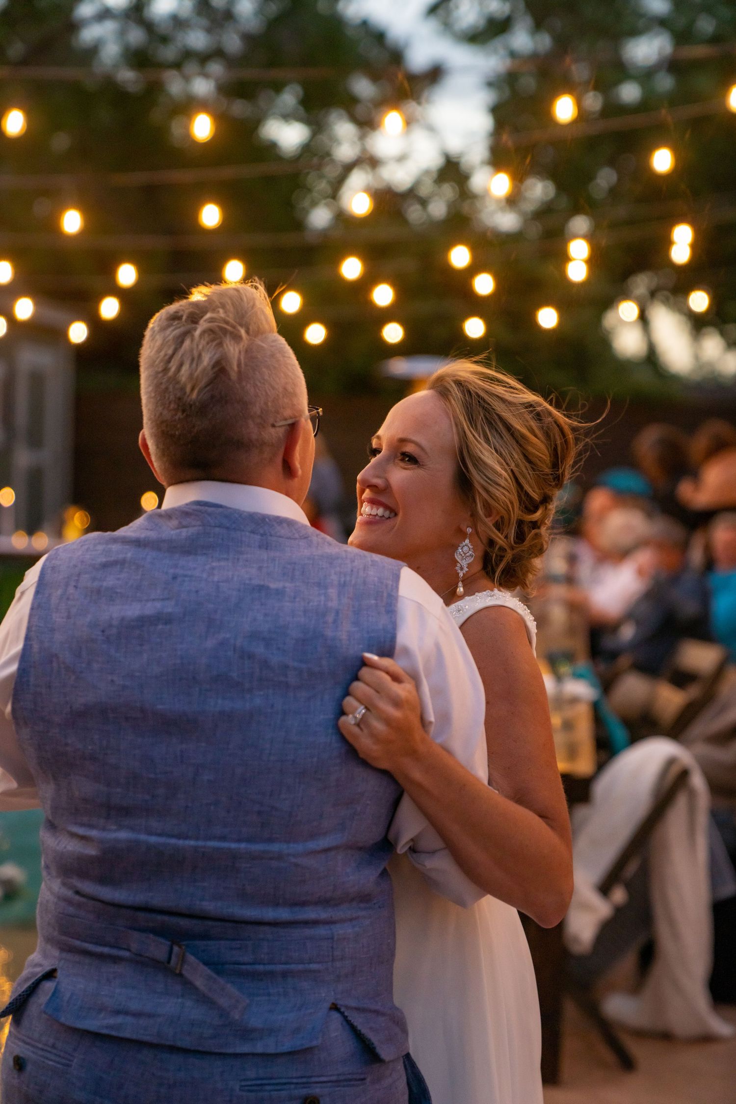 A couple shares a romantic dance under twinkling string lights at an outdoor evening wedding reception.