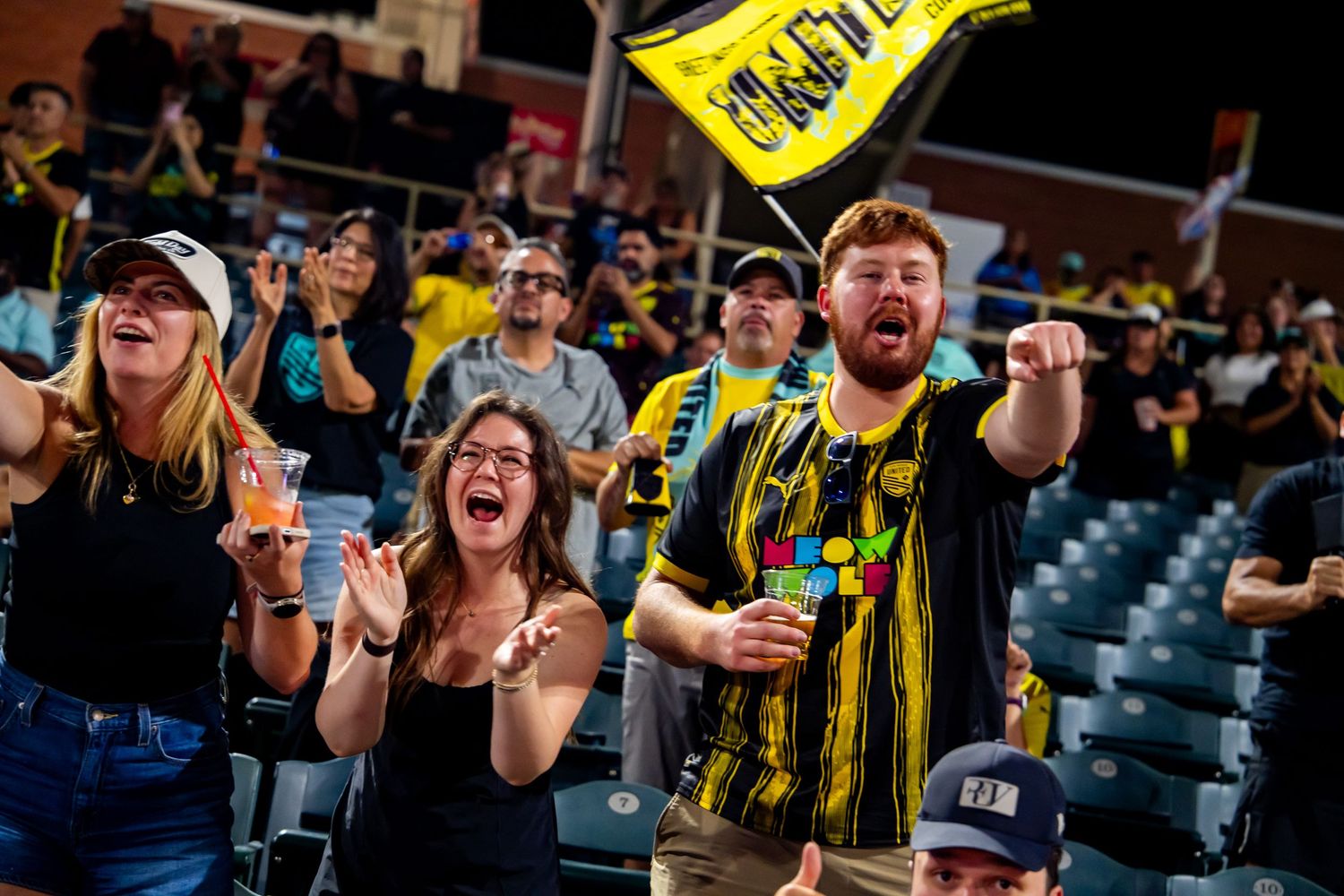 Excited soccer fans in black and yellow jerseys cheering enthusiastically at a night game in a stadium.