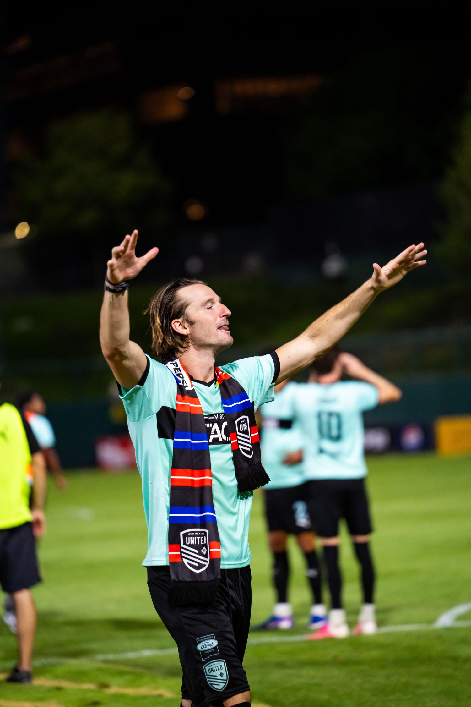 Soccer player in teal jersey celebrates with arms raised, wearing colorful scarf on field at night.