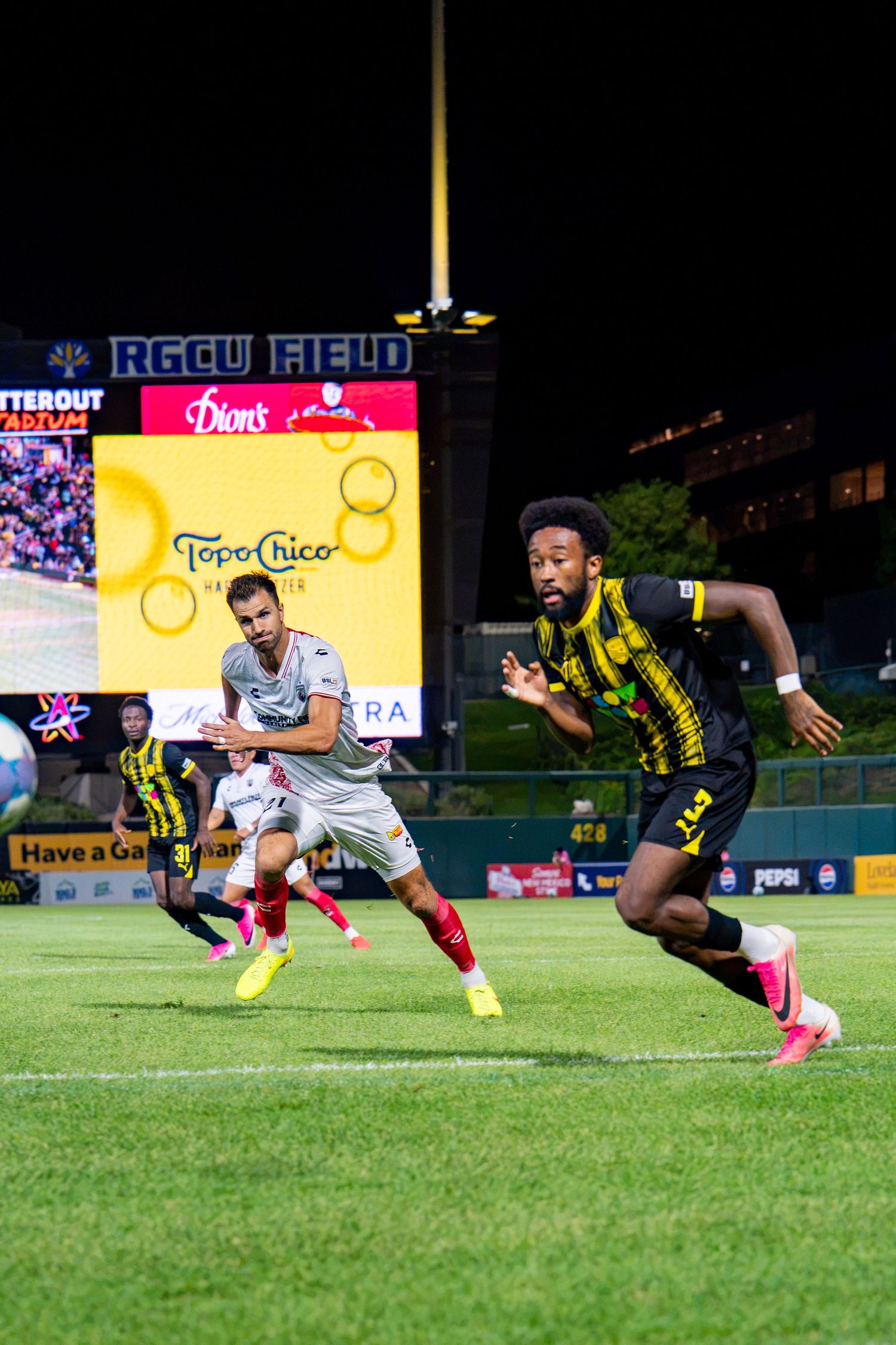 Two soccer players compete at night on RGCU Field, with Topo Chico and Dion's advertisements visible.