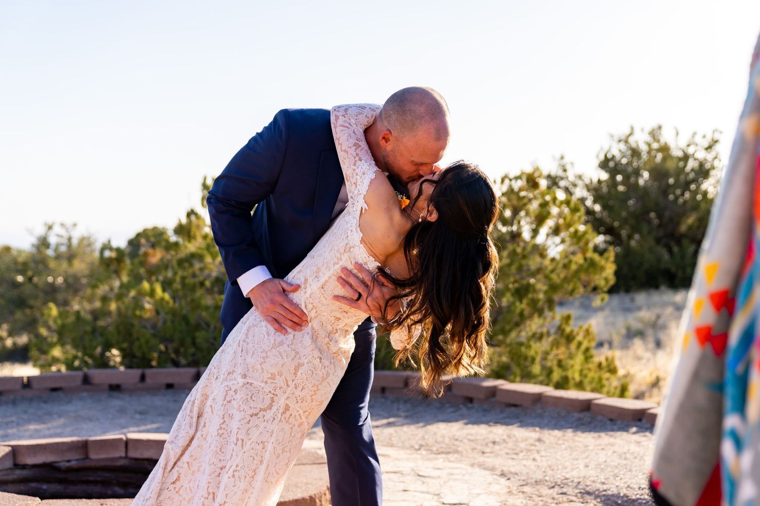 A groom in a navy suit dips his bride in a lace wedding dress for a romantic kiss outdoors.