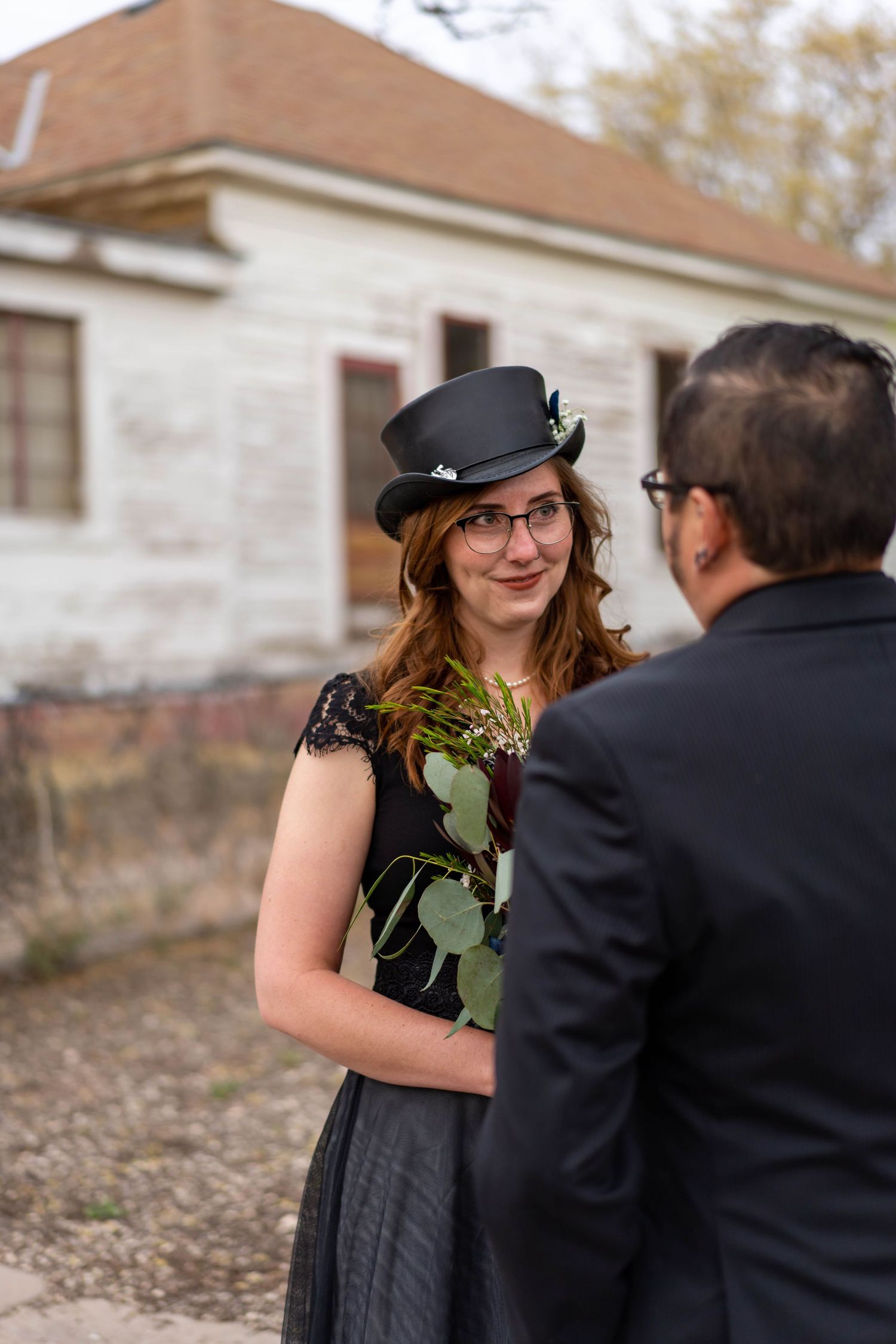 A couple shares a romantic moment outdoors, with one wearing a vintage-style black hat and dress near a stone building.