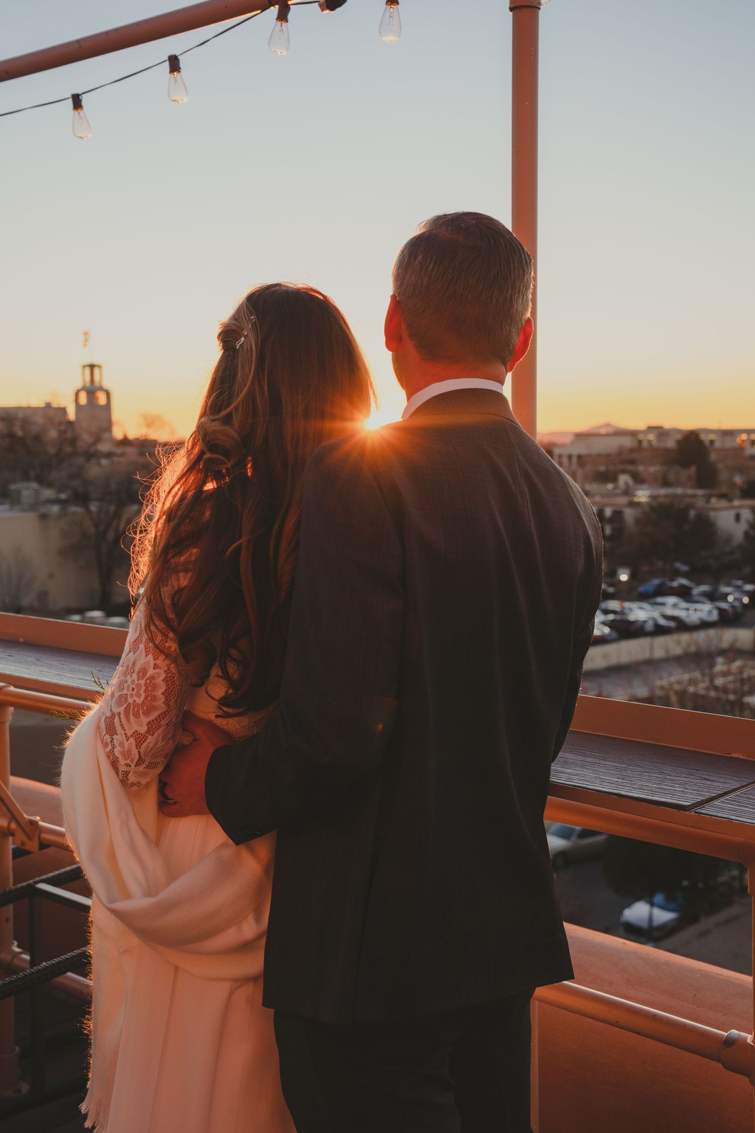 Silhouetted couple watches sunset from a rooftop terrace with string lights overhead.