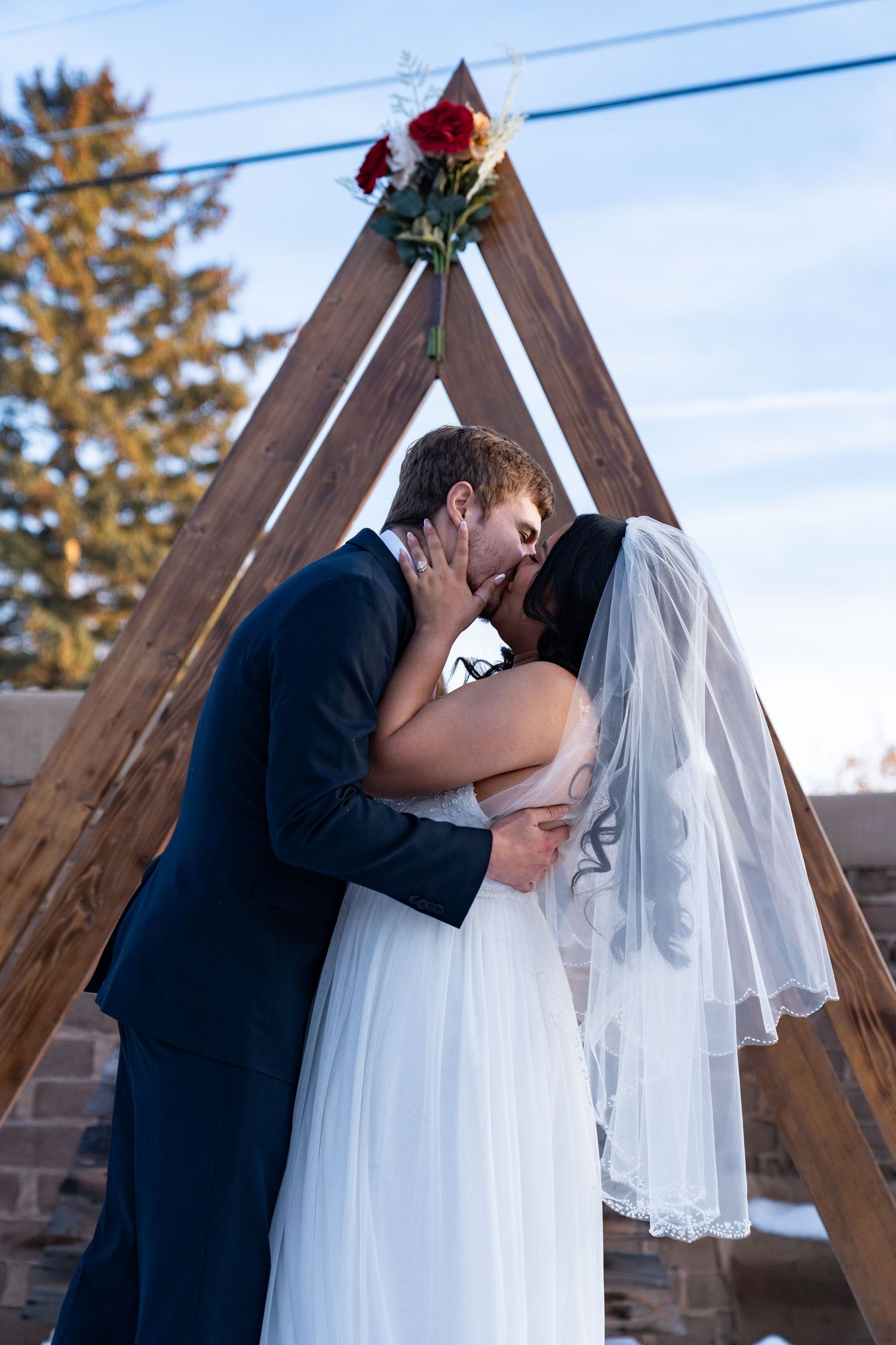 A romantic wedding ceremony moment under a wooden arch decorated with a red rose against a blue sky.