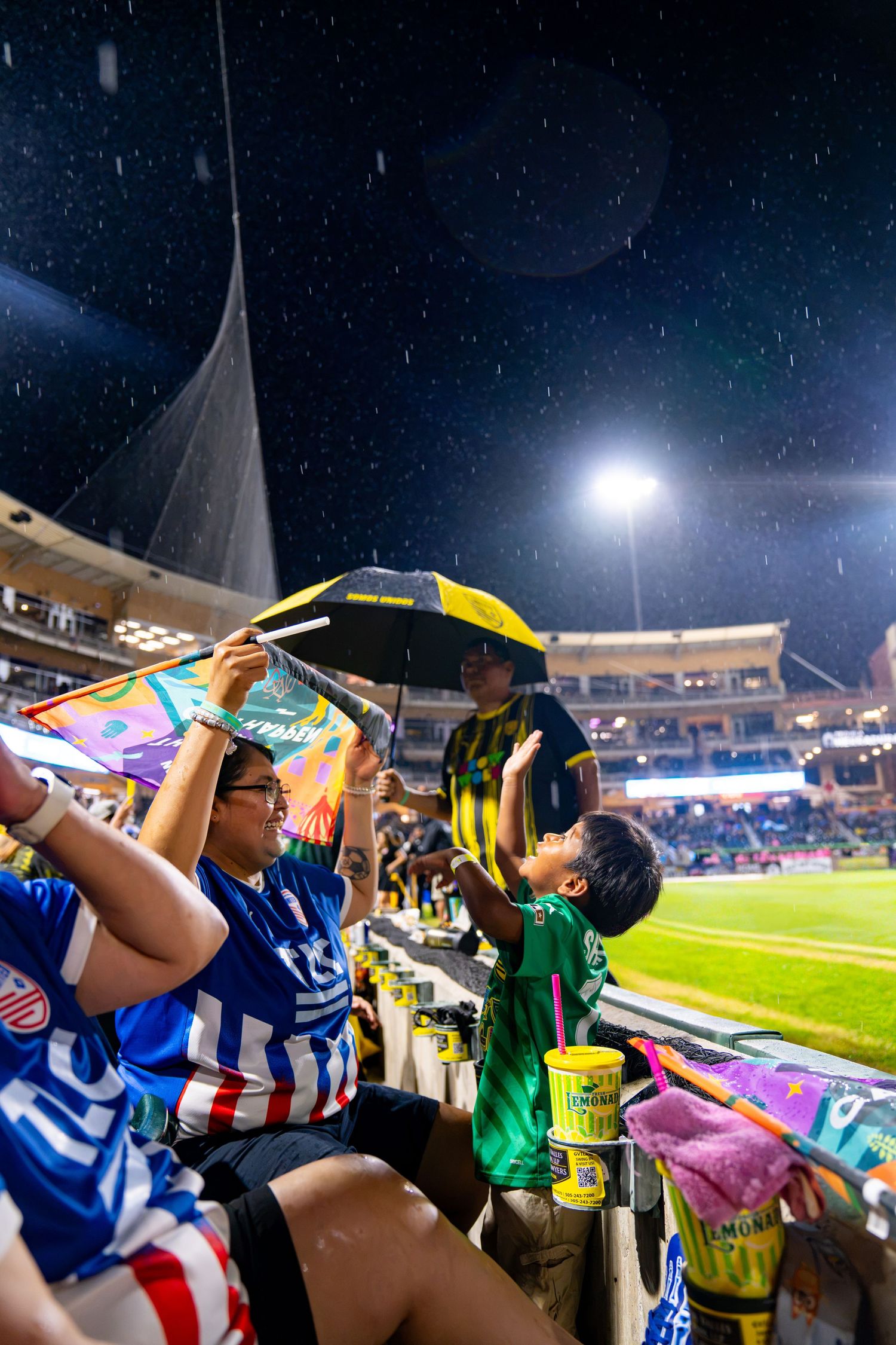 Fans shelter from rain with umbrellas in stadium stands during a night soccer match under bright floodlights.