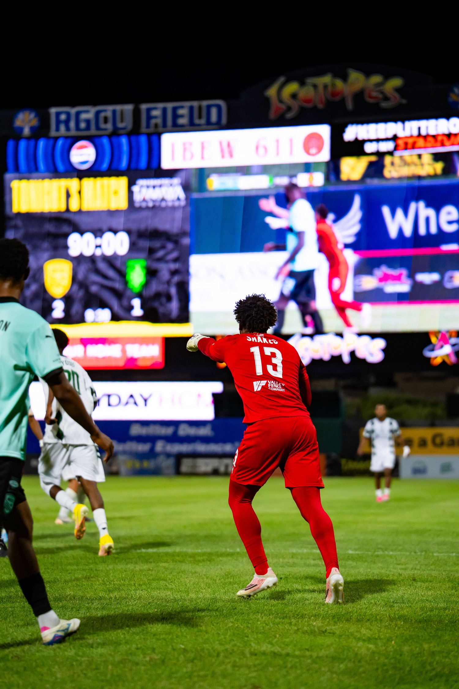 Soccer player #13 in red jersey plays at RGCU Field stadium at night with scoreboard lit up behind.