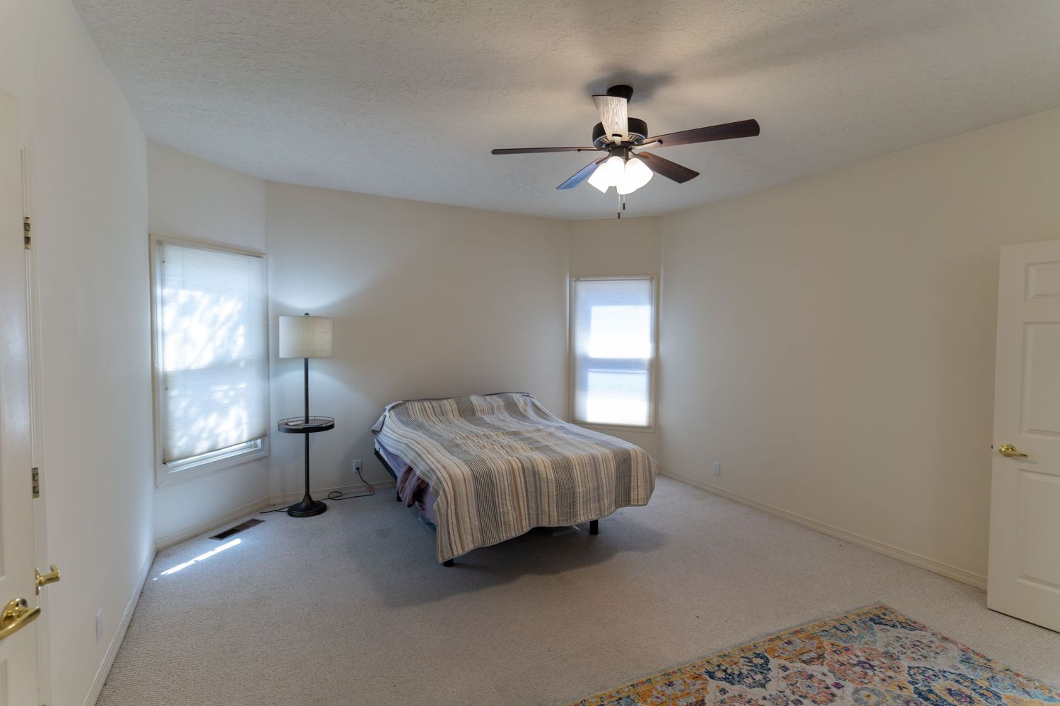 Vacant room with beige carpet, white walls, and ceiling fan illuminated by natural light from windows.