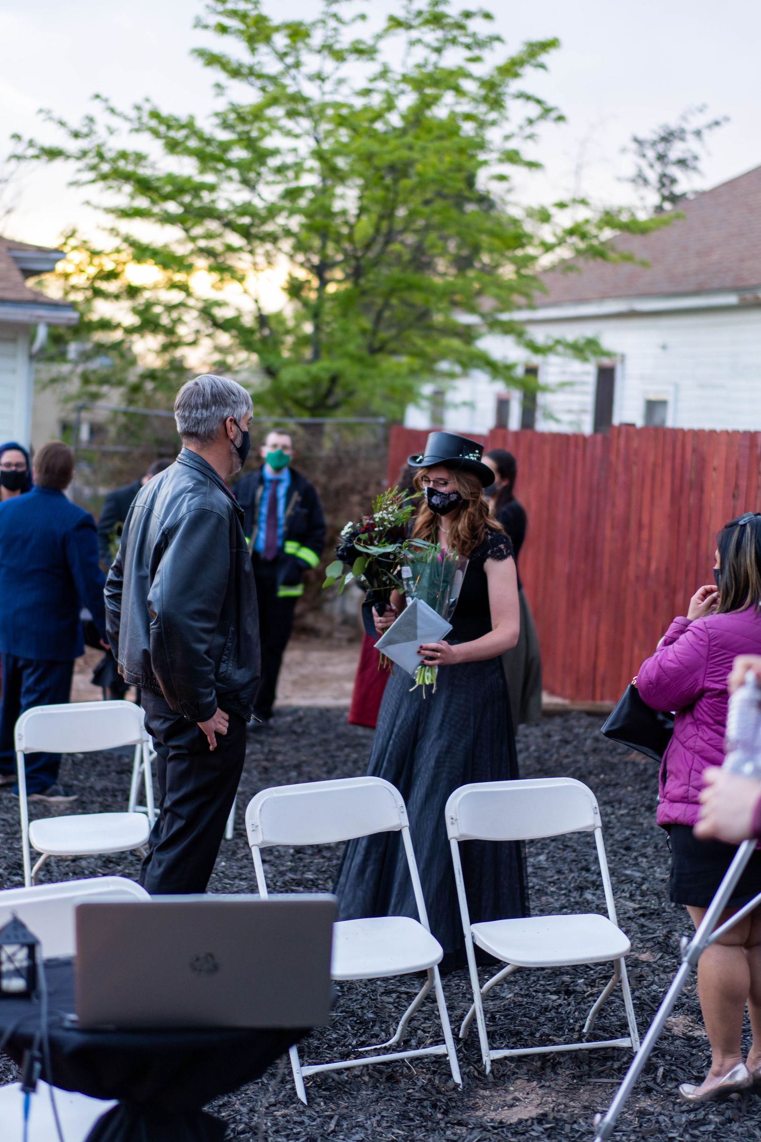 People in masks gather outdoors around white folding chairs during a socially distanced event.