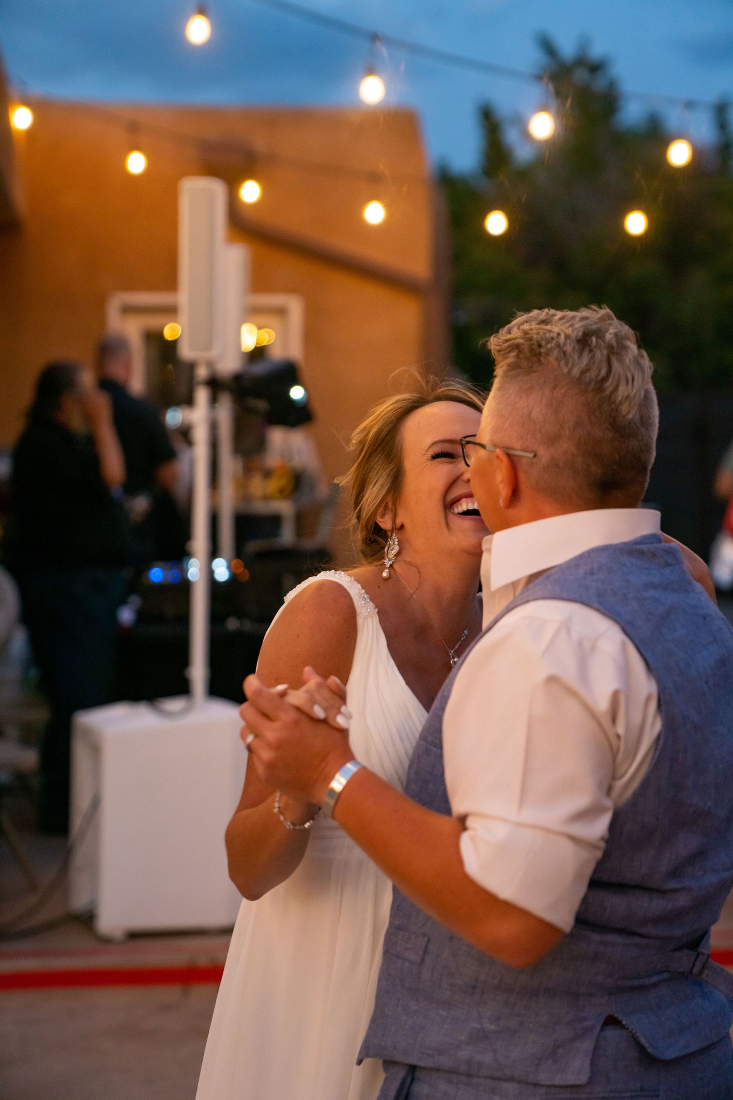 A couple dances and laughs together at their evening wedding reception under glowing string lights.