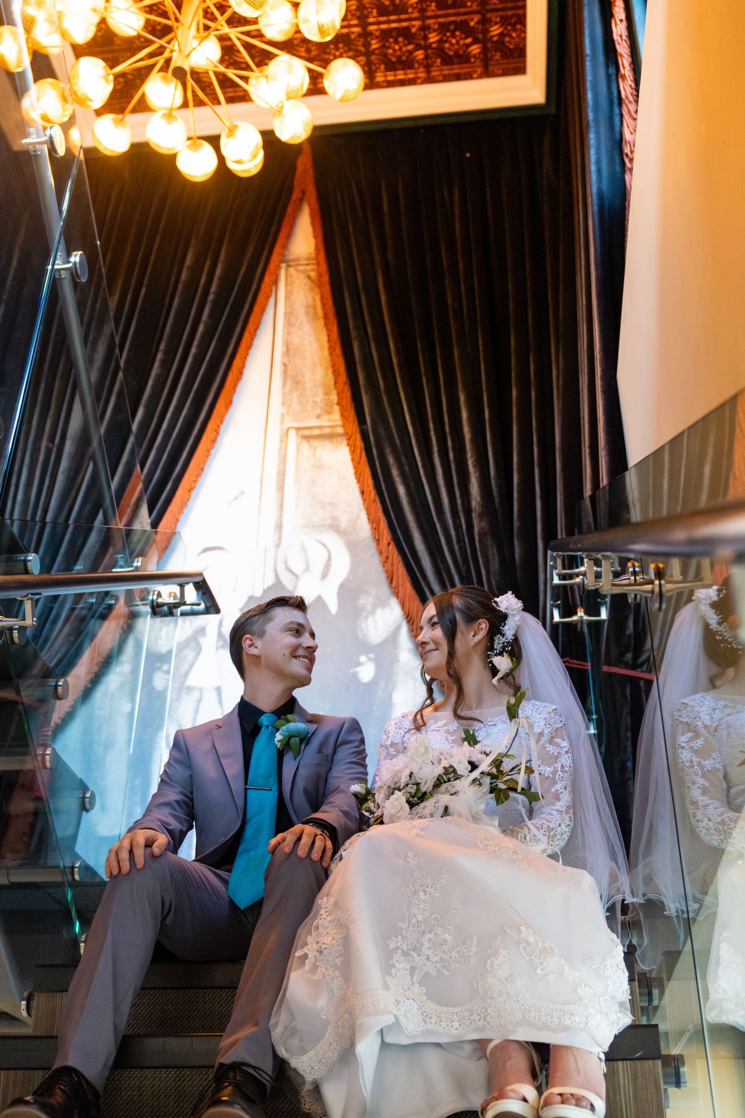 Bride and groom sitting on stairs, smiling at each other, chandelier above, dark curtains background.