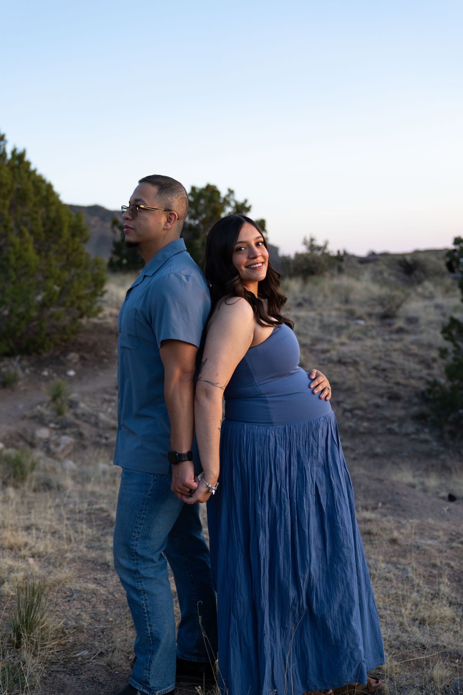 Couple standing back-to-back outdoors at dusk, woman in blue maxi dress cradling baby bump, holding hands in desert landscape.