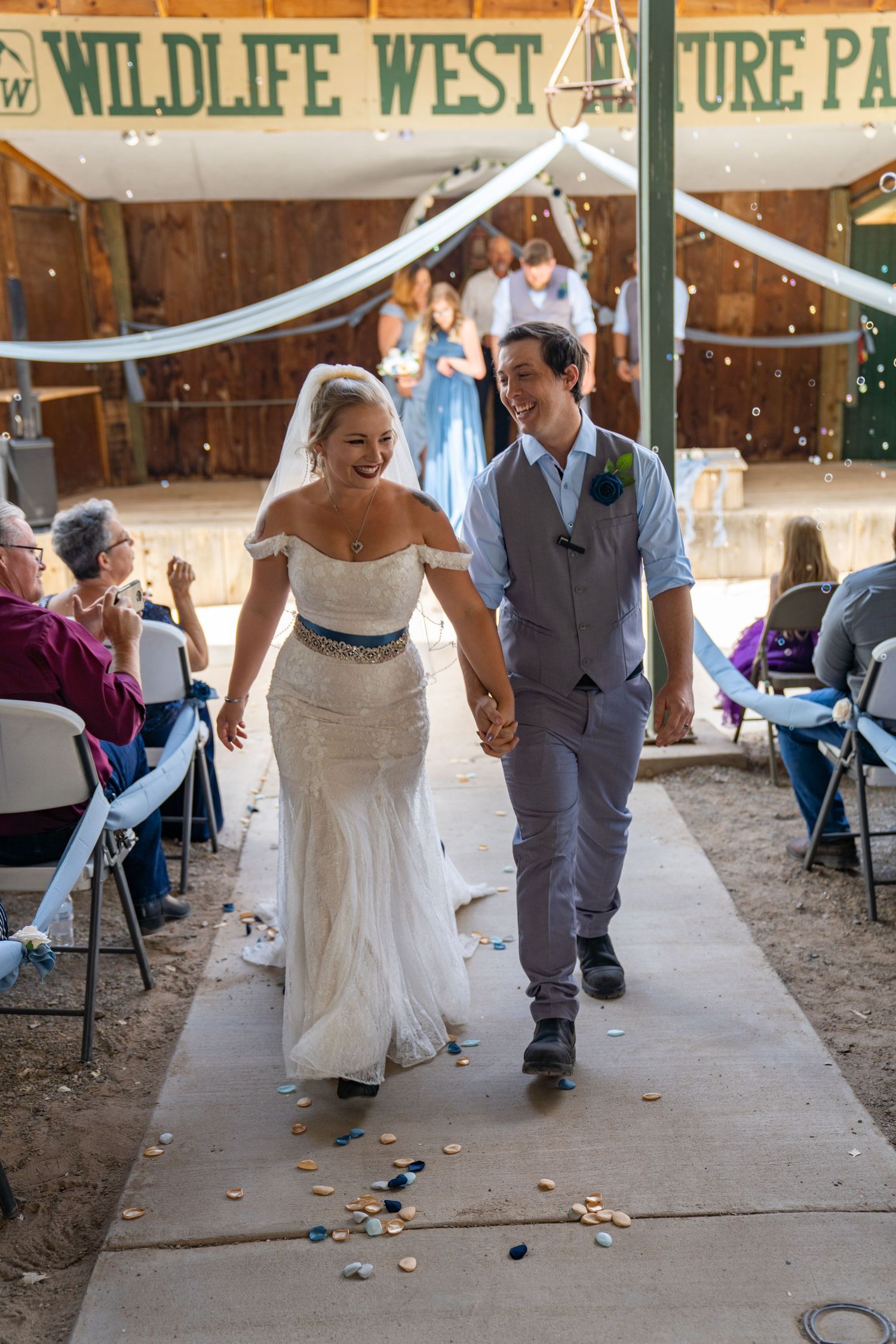 Wedding couple playfully walks down aisle while tossing flower petals in rustic barn venue.