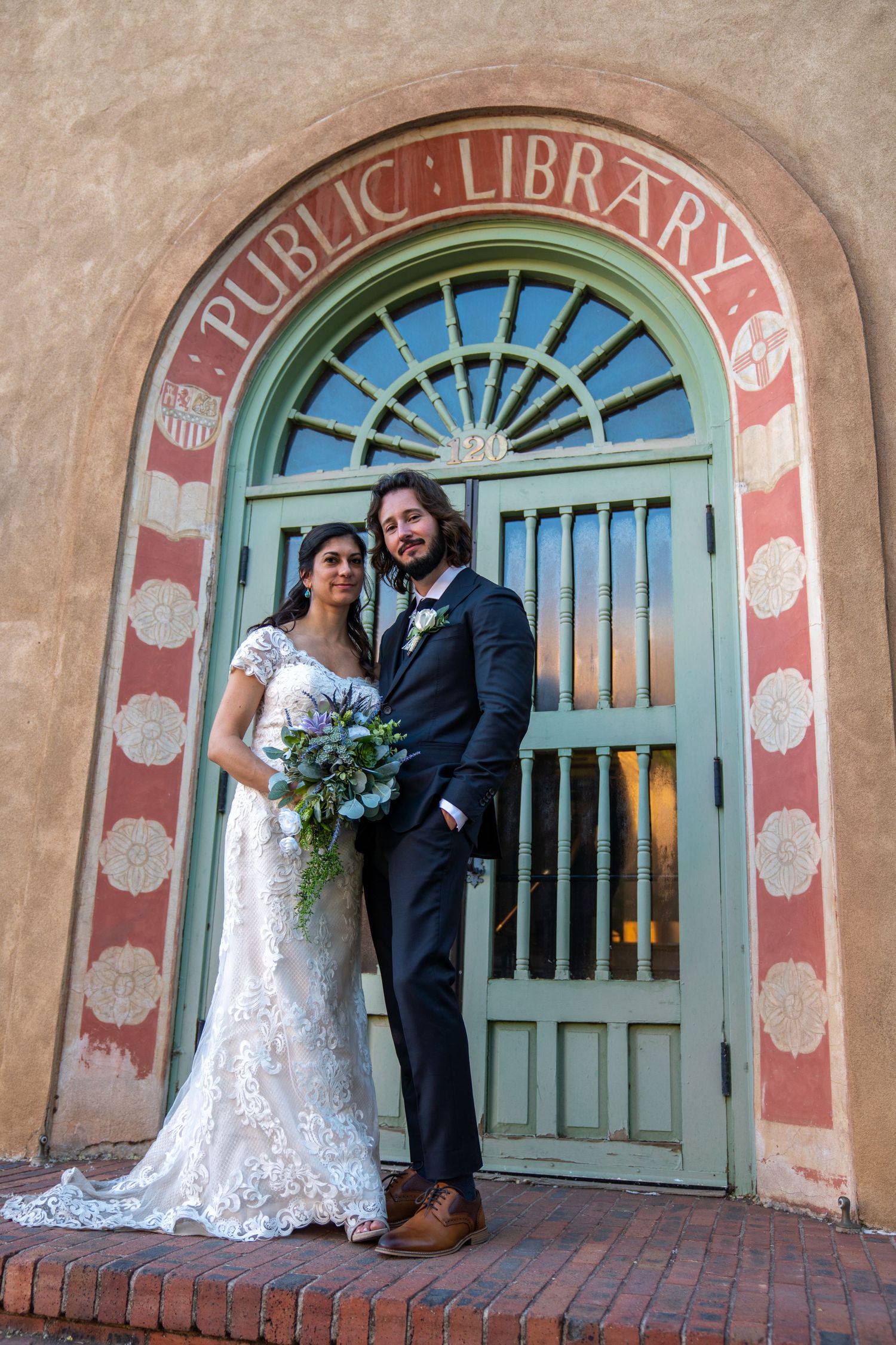 A couple poses in formal wedding attire in front of a vintage public library entrance with turquoise door.