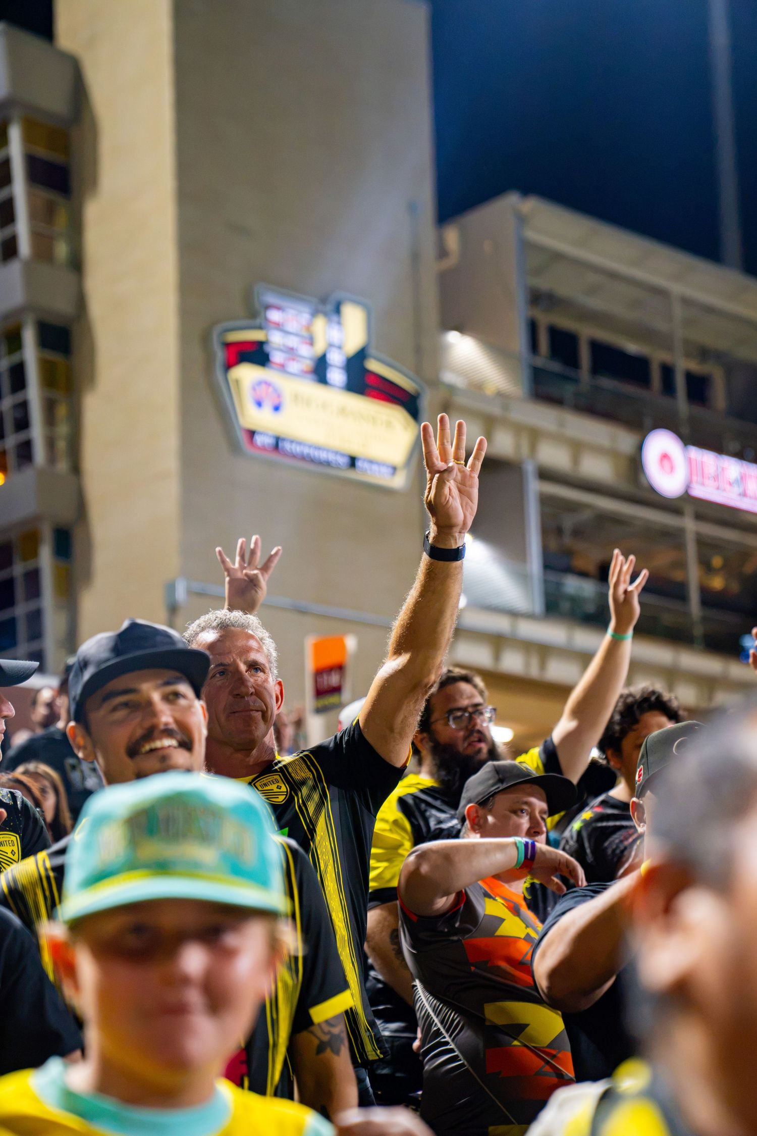 New Mexico United soccer fans celebrating with raised hands at a night game wearing black and yellow team colors.