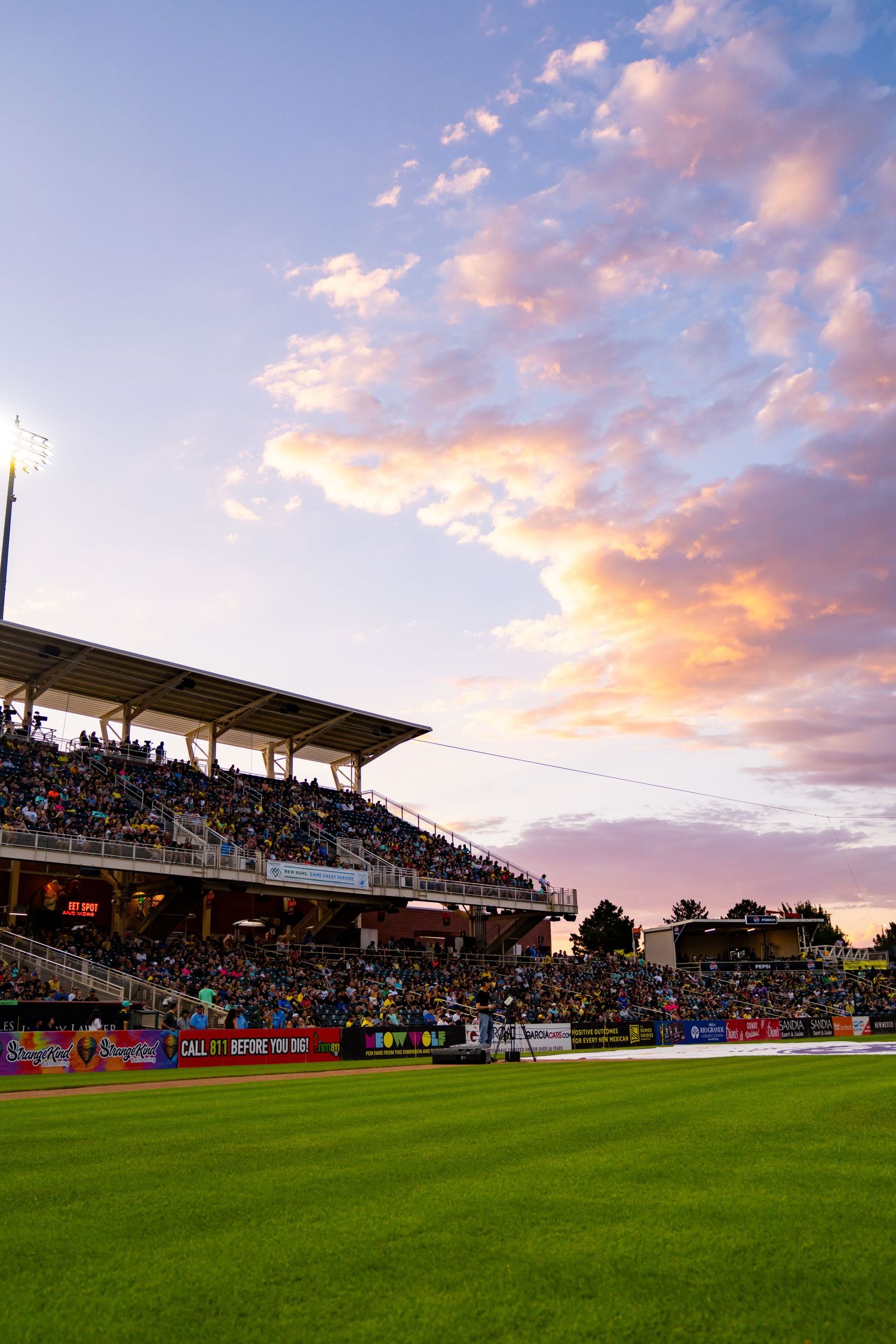 Packed stadium at sunset with vibrant pink and orange clouds over a lush green field during an evening sports event.