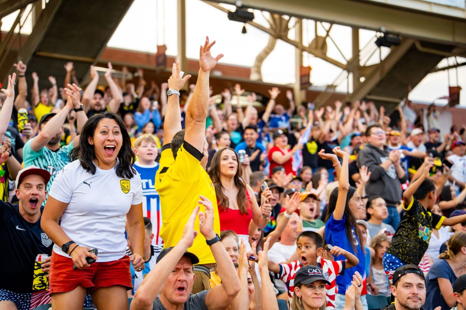 Excited soccer fans celebrate in the stands, cheering with arms raised at an outdoor stadium during a match.