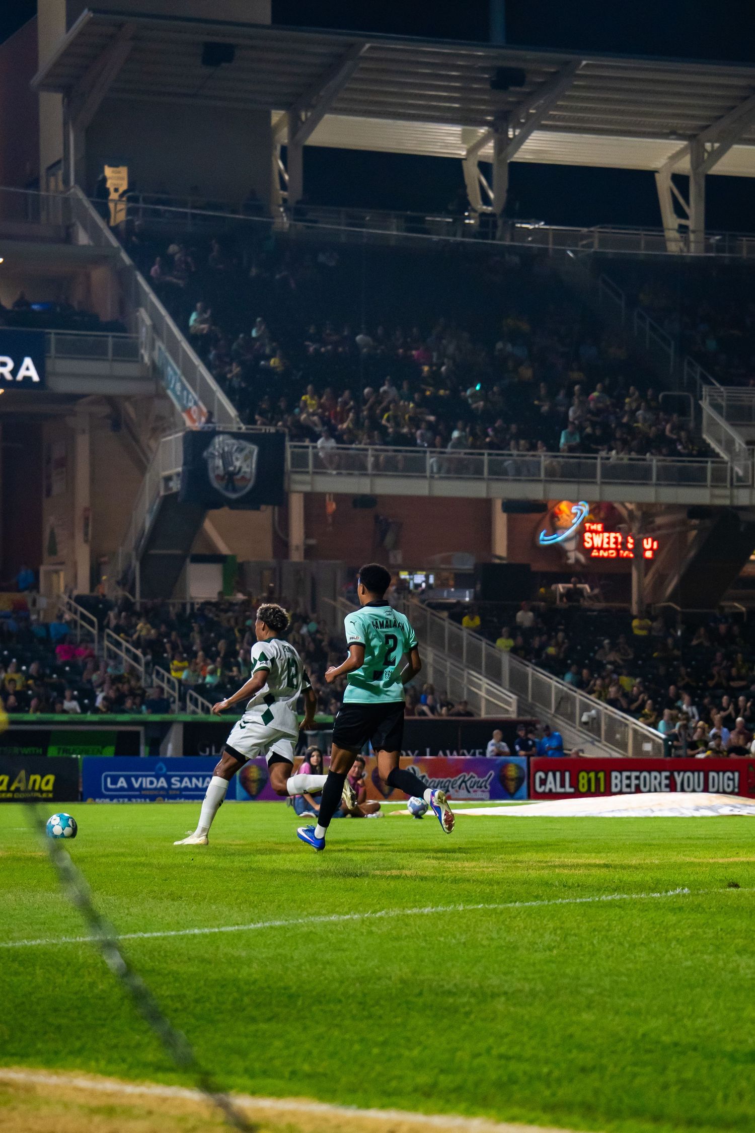 Soccer players compete on a floodlit stadium pitch at night with crowds in the stands during a professional match.