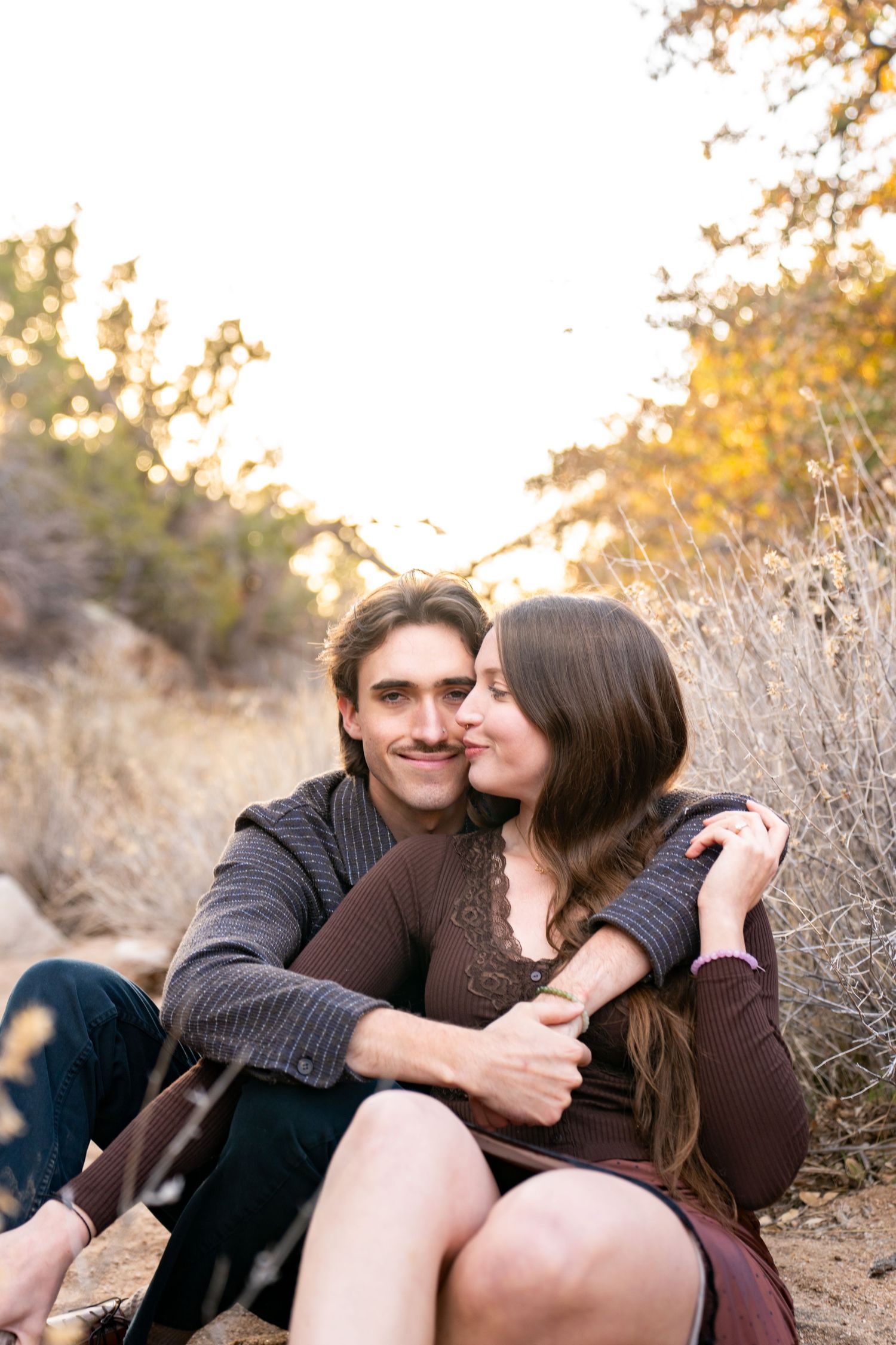 Couple sitting outdoors in golden light, woman kissing man on cheek while embracing among dry brush.