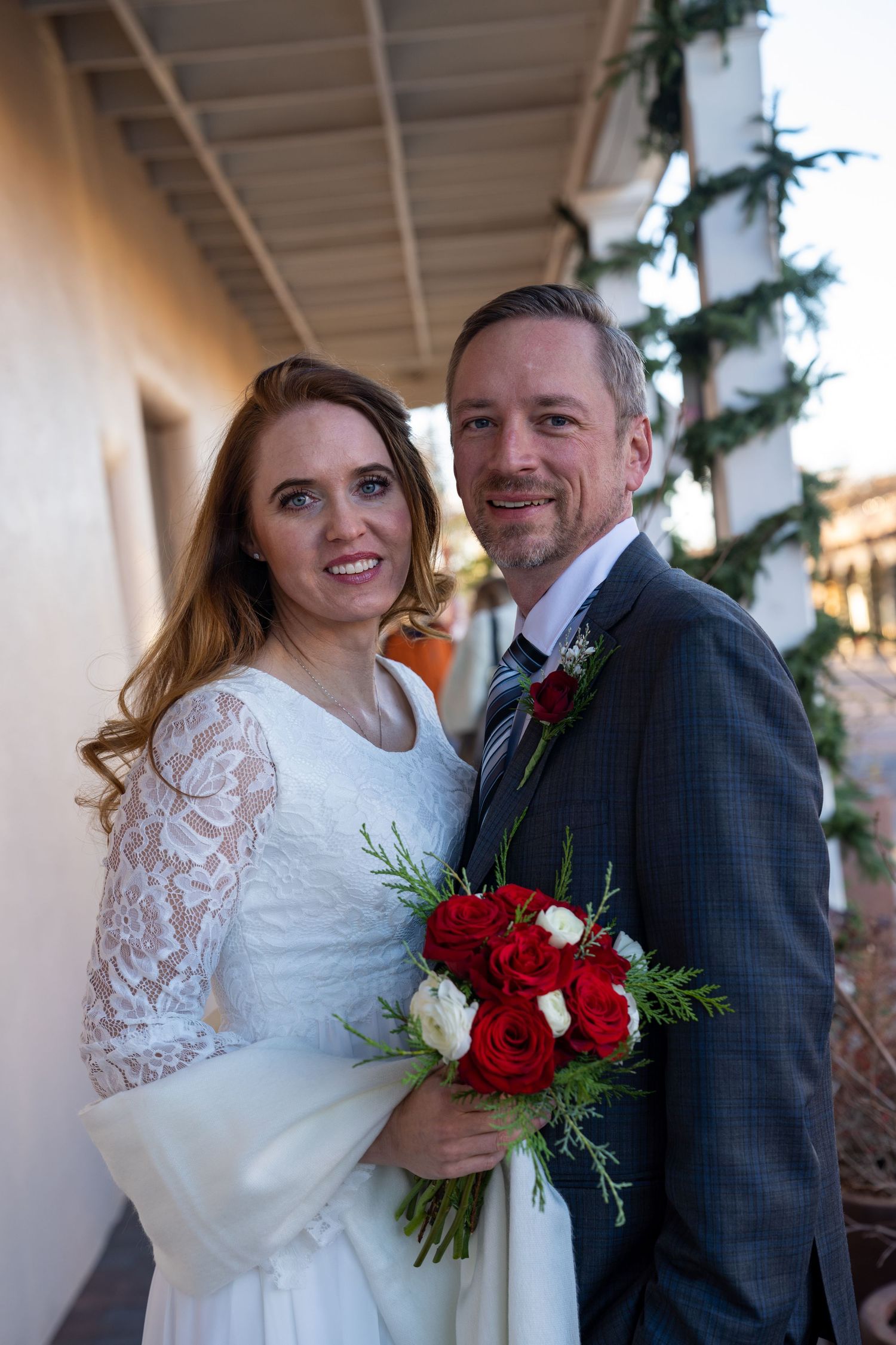 Newlywed couple shares intimate moments in a hallway decorated with holiday greenery and red rose bouquet.