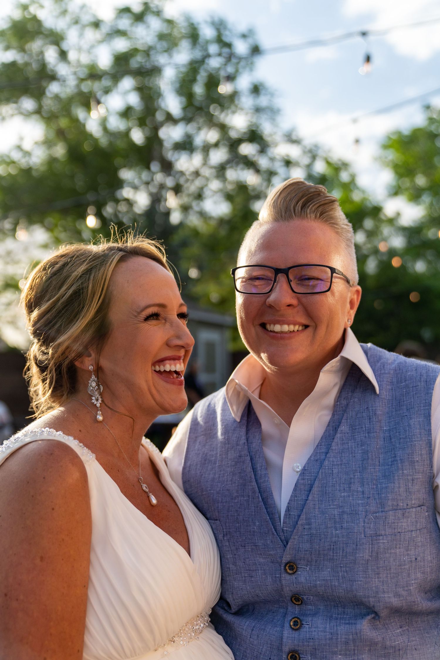Couple smiles and embraces during sunset wedding celebration with string lights and greenery.