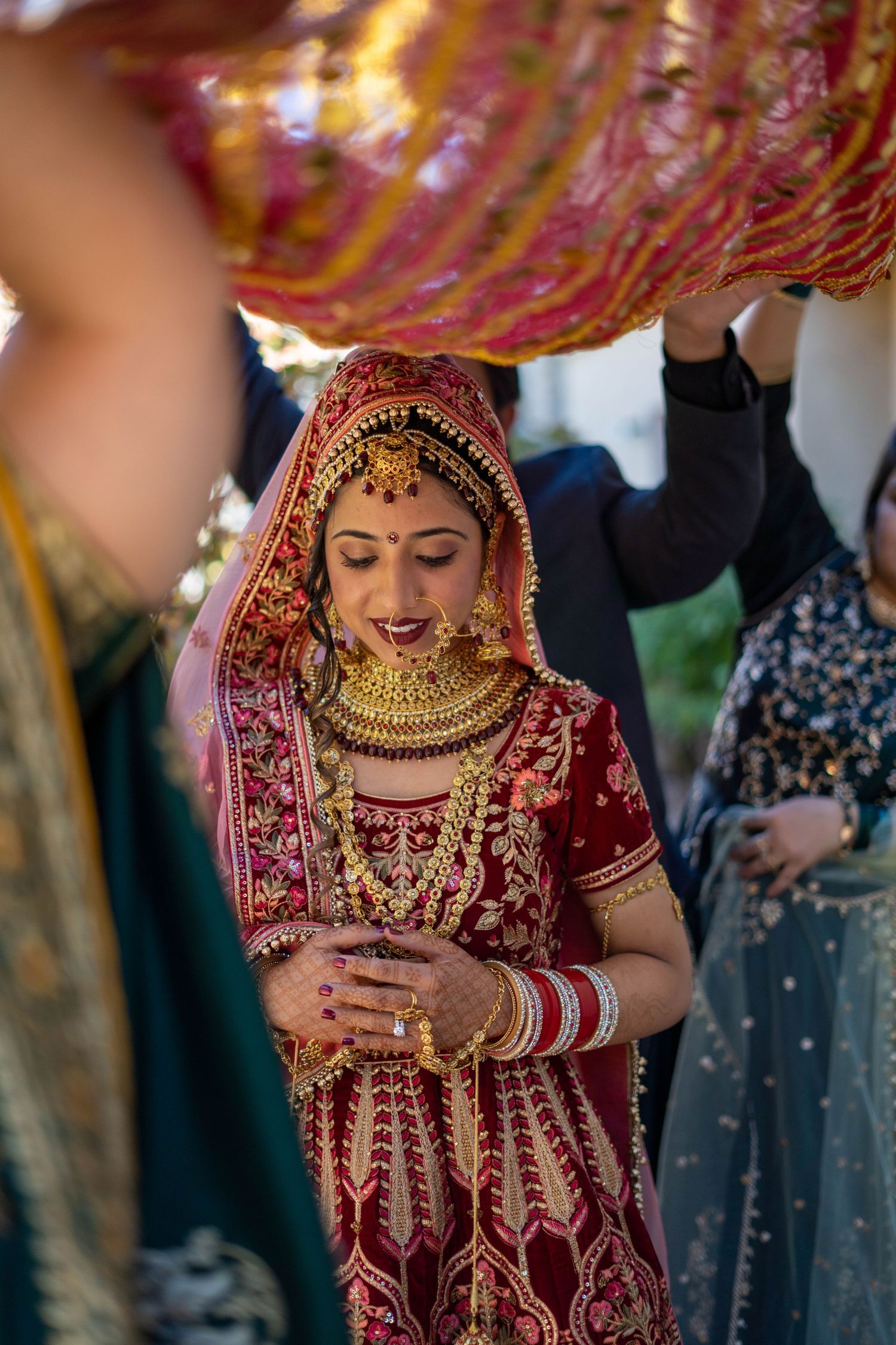 A bride in a red and gold lehenga with traditional jewelry stands under decorative balloons.
