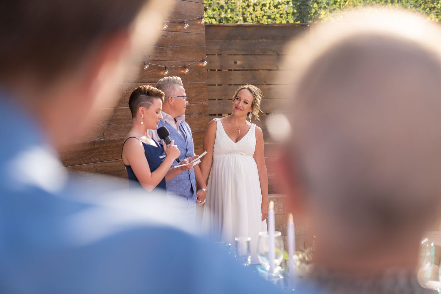 People share a joyful moment during an outdoor wedding reception with candlelit decor and a wooden backdrop.