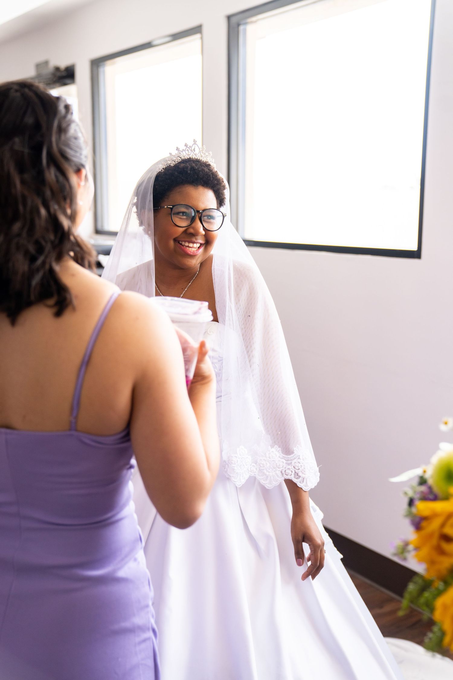 A bride in a white wedding dress and glasses smiles while talking with someone wearing a purple bridesmaid dress.