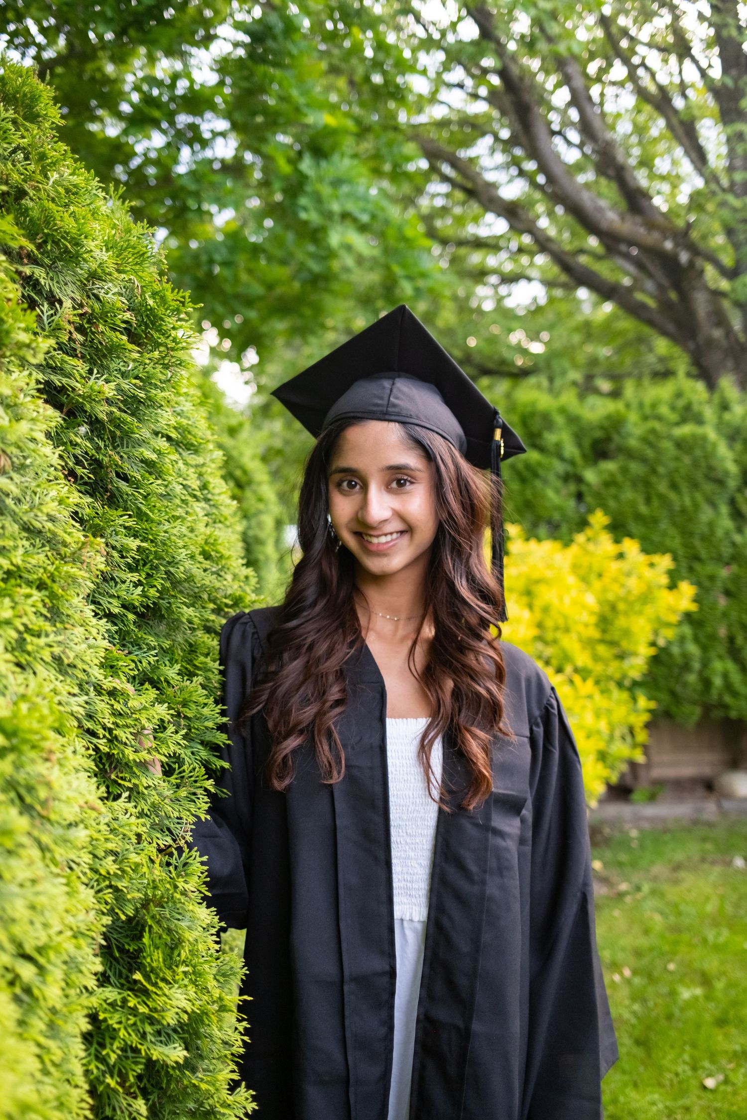 Graduate in cap and gown poses in front of green foliage.