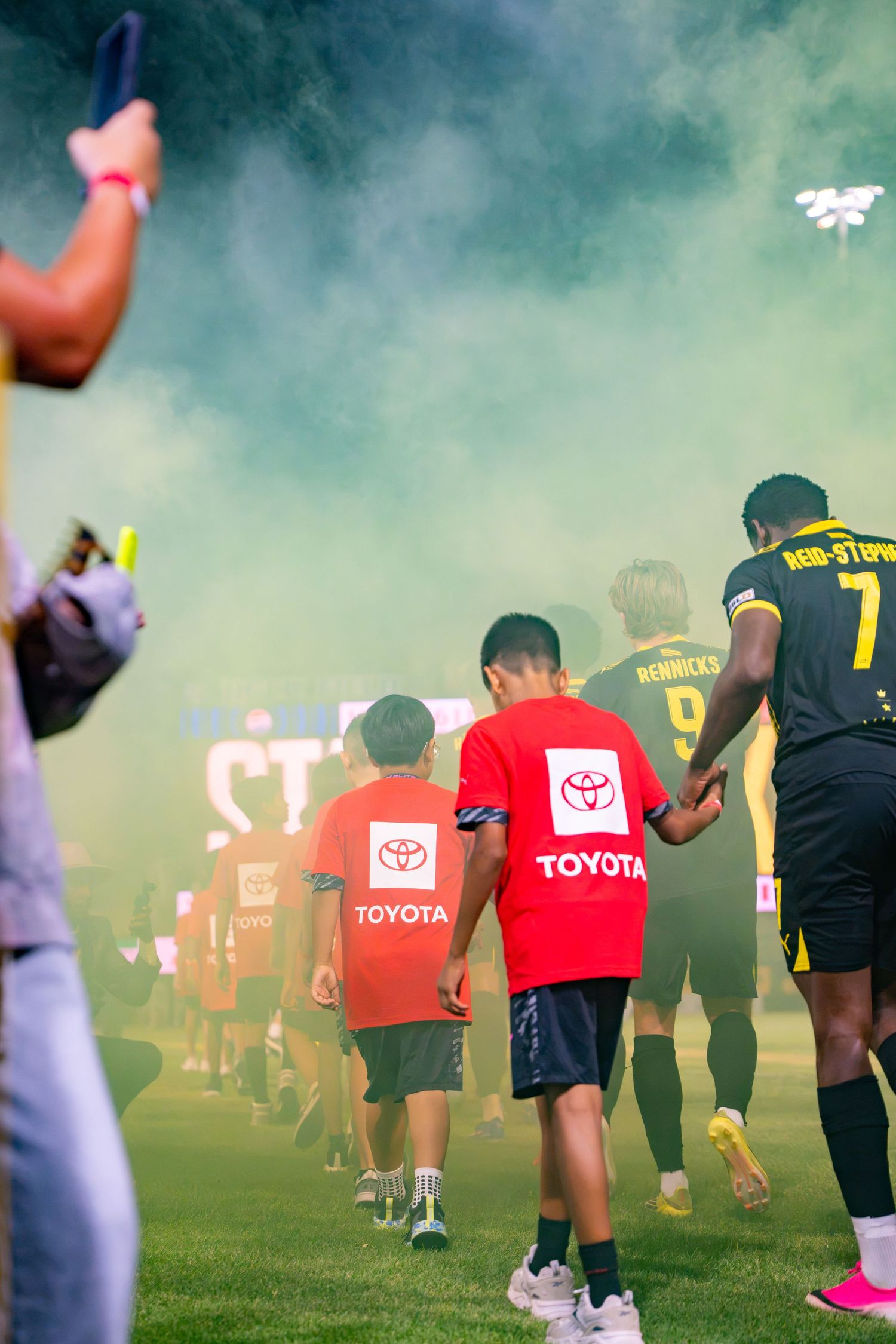 Kids in Toyota shirts walk with soccer players through green smoke at a stadium pre-game ceremony.