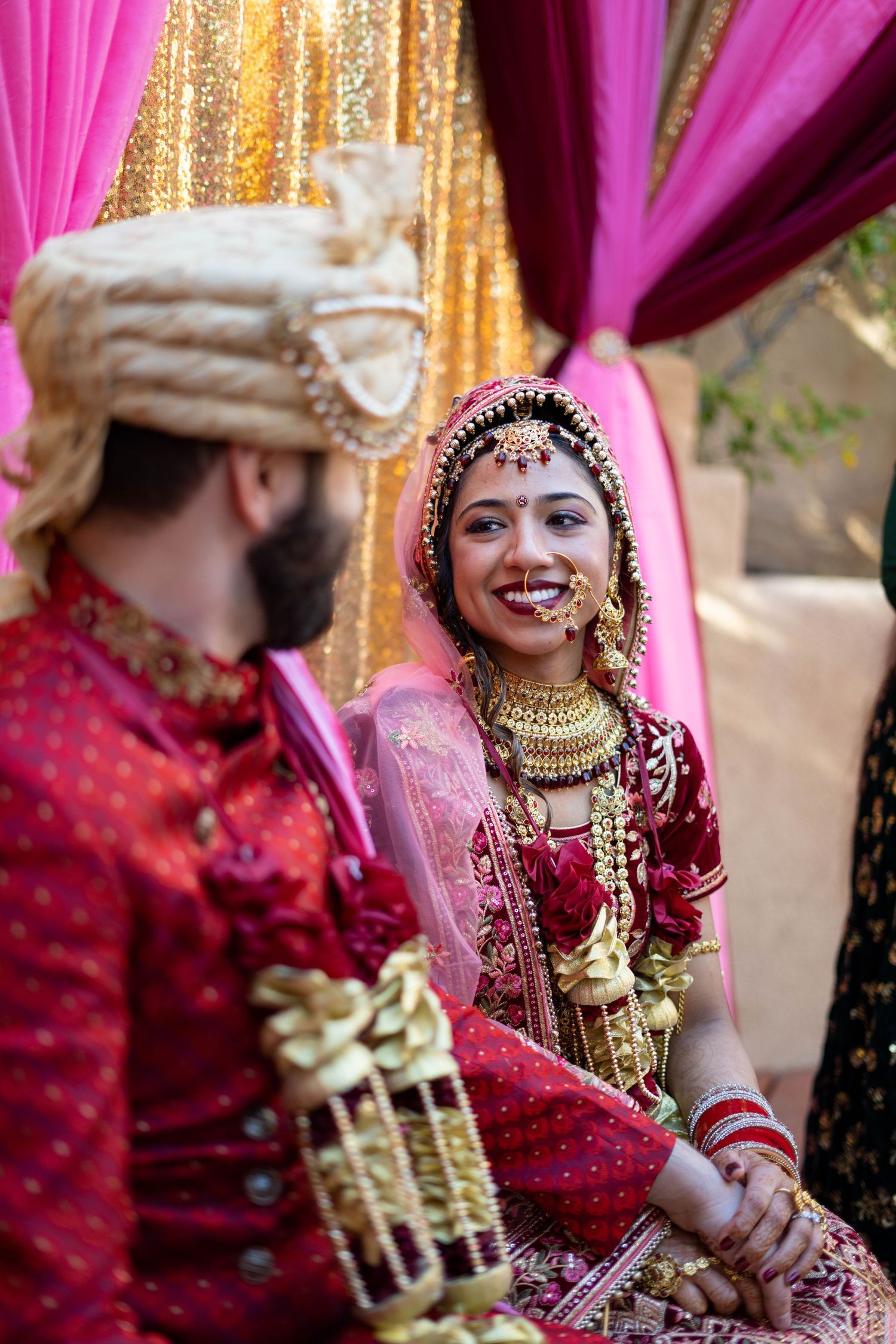 Traditional Indian bride wearing red and pink wedding attire with ornate gold jewelry smiles during ceremony.