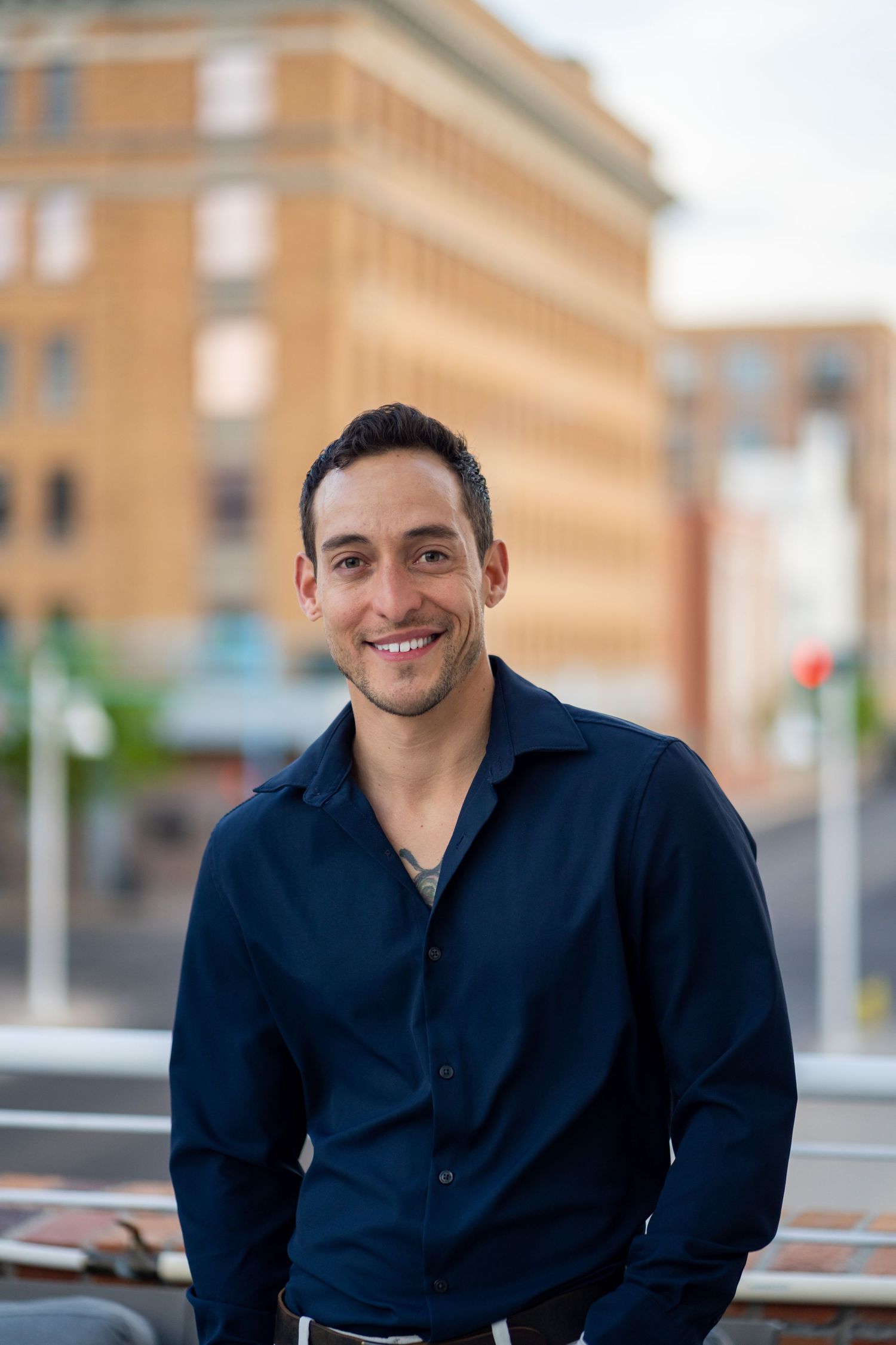 Professional portrait of a person in a navy button-down shirt smiling in an urban setting.