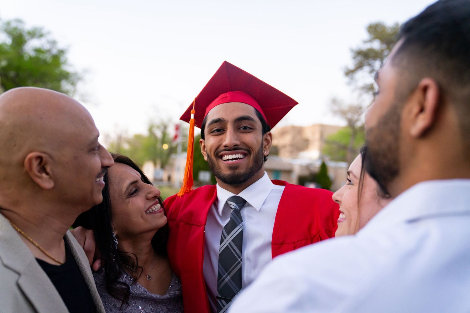 Smiling graduate in red cap and gown celebrates with proud family members outdoors at graduation ceremony.