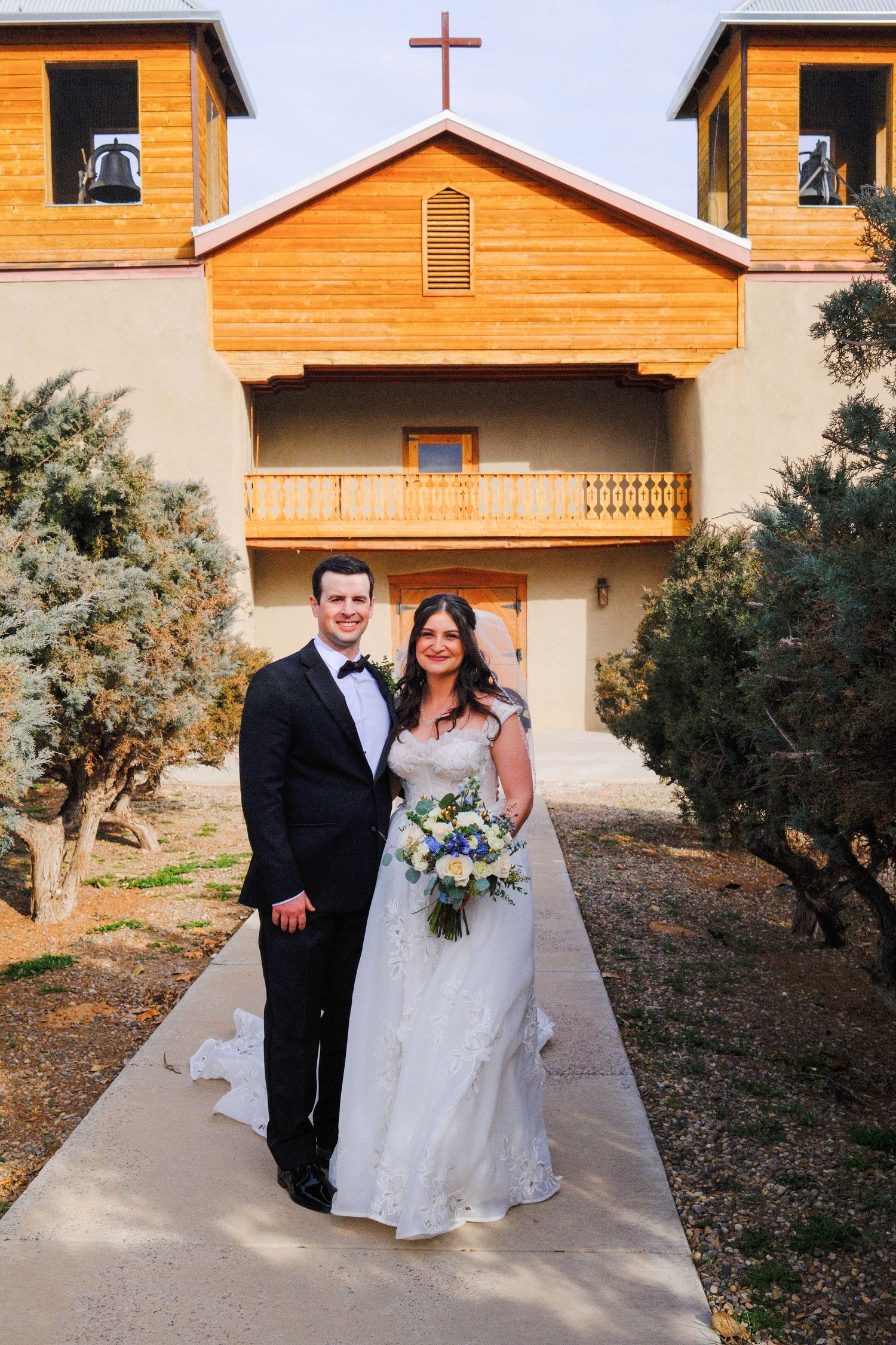 Bride and groom posing outside a rustic church with a cross, bride holding a blue and white bouquet.