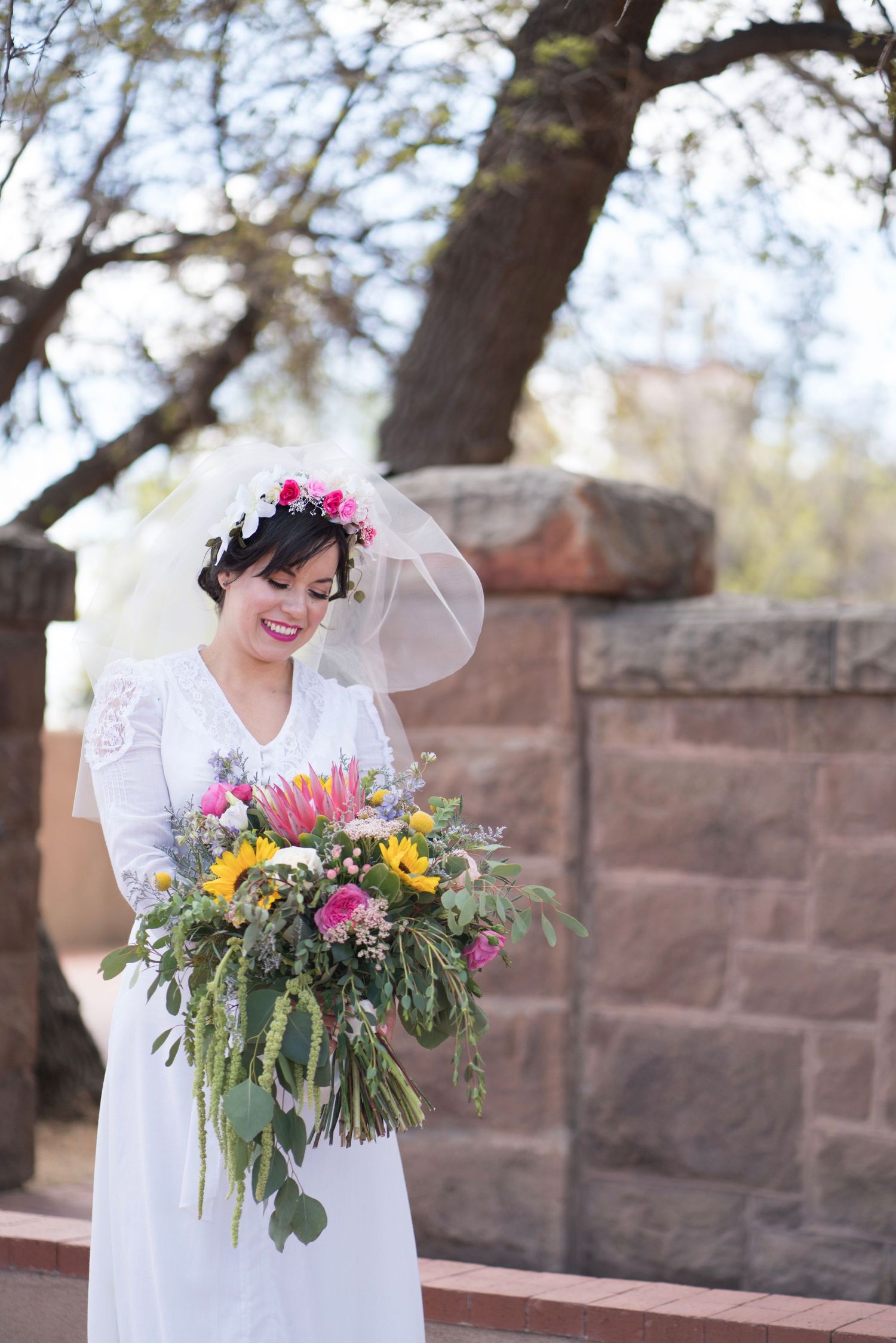A bride in a white dress holds a vibrant bouquet with pink and yellow flowers against a stone wall backdrop.