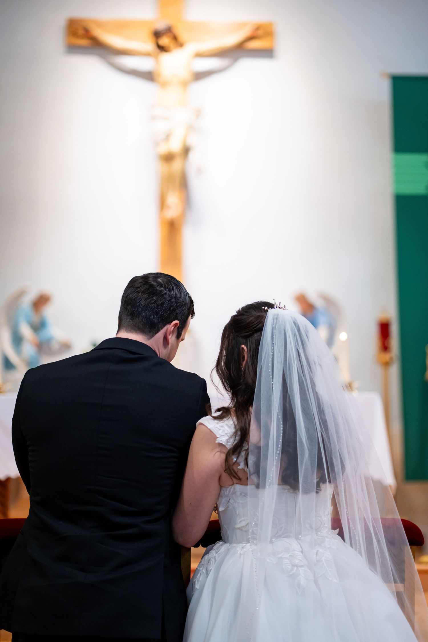 Bride and groom kneeling together in prayer before a crucifix during their Catholic church wedding ceremony.