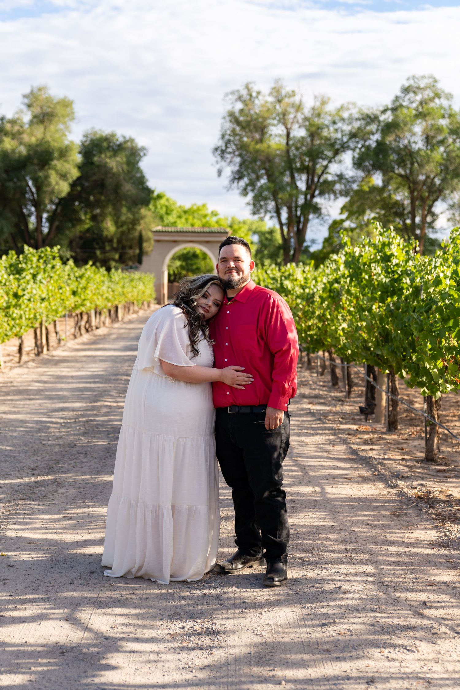 A couple poses together in a vineyard with rows of grapevines.